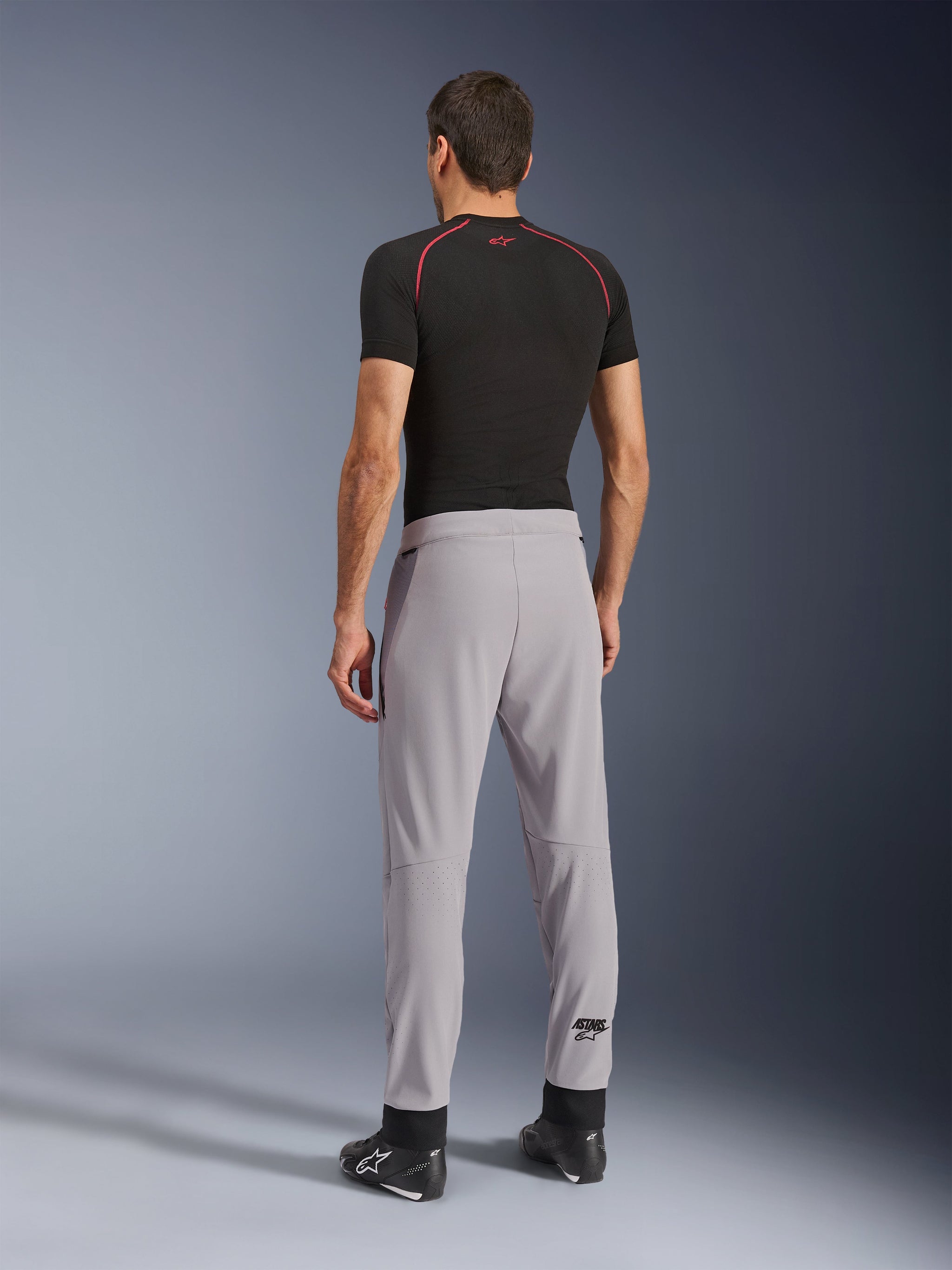 Achteraanzicht van een model dat de Alpinestars Aeroshell Airflow broek in Mid Gray draagt, gecombineerd met een zwart basislaag shirt en zwarte schoenen, waarbij de laser-geperforeerde zones op de kuiten en de sportieve pasvorm worden benadrukt.