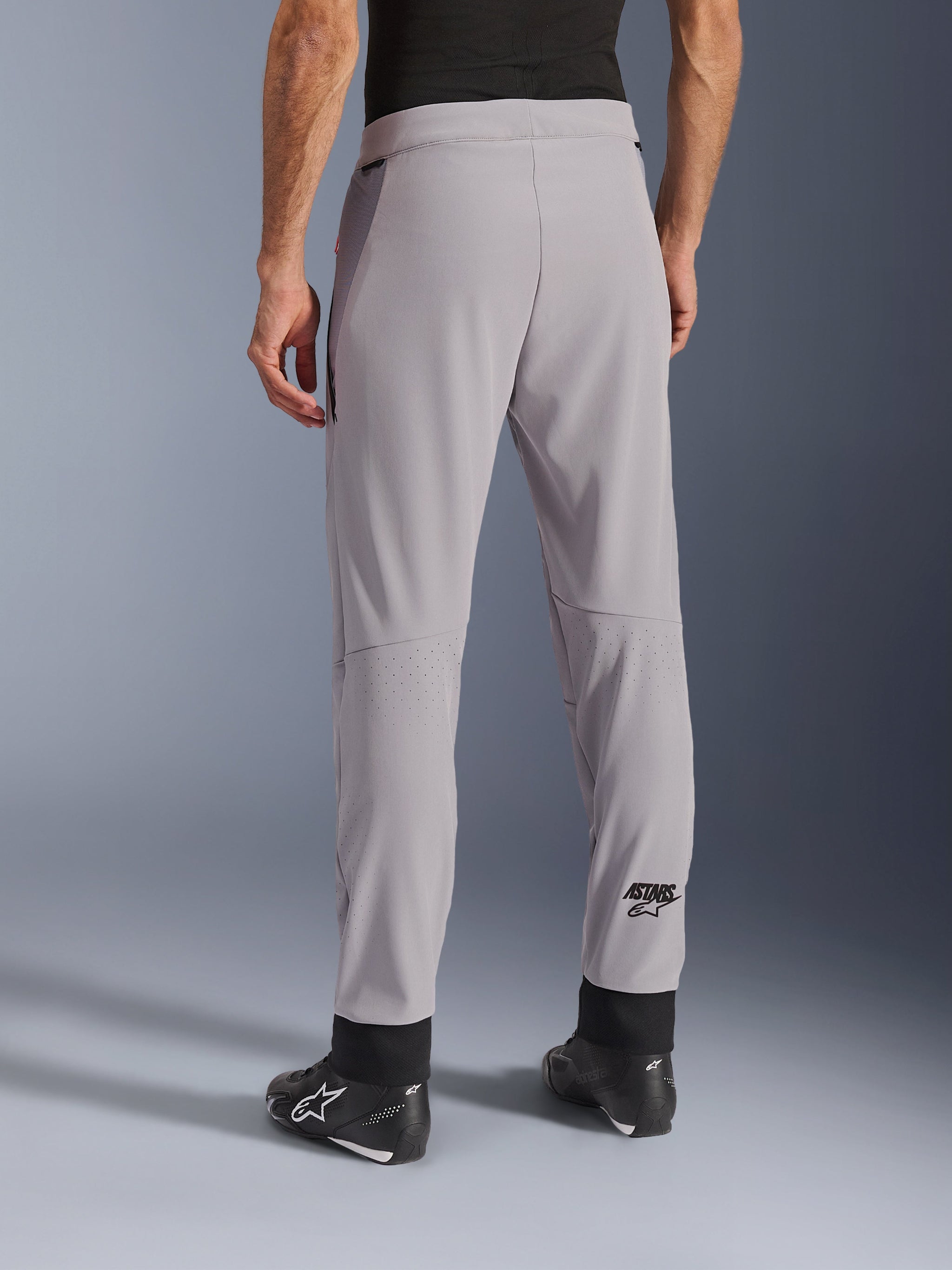 Alpinestars Aeroshell Airflow broek, Mid Gray, achteraanzicht met laser-geperforeerde zones op de kuiten, ASTARS-logo op het onderbeen, zwarte verstelbare enkelmanchetten, gedragen met zwarte motorlaarzen, lichtgewicht sportieve textiele motorbroek