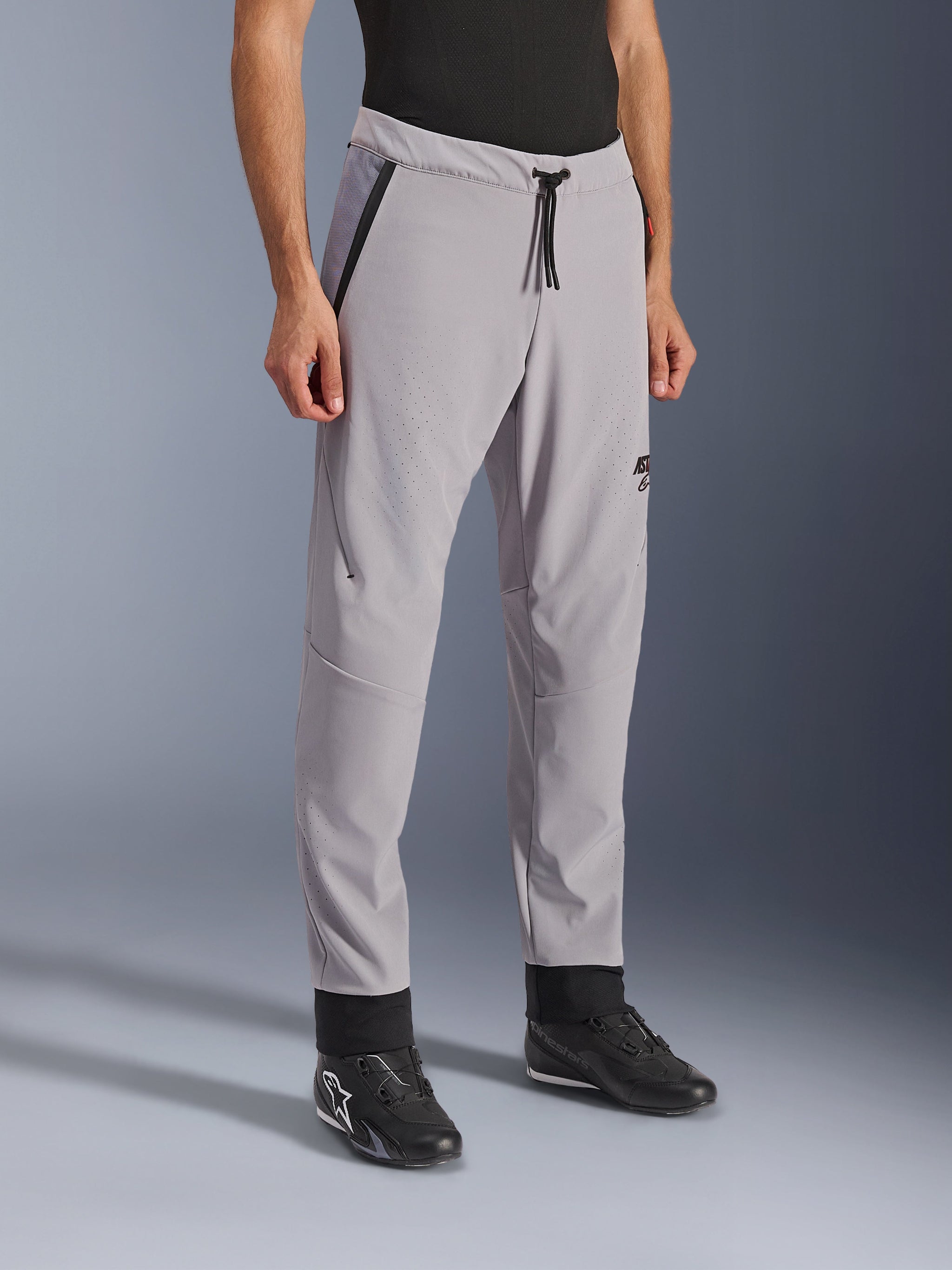 Alpinestars Aeroshell Airflow Broek, Mid Gray, lichtgewicht motorbroek met laser-geperforeerde ventilatiezones op de dijen en schenen, voorzien van een elastische tailleband met trekkoord en een slanke sportieve pasvorm, gedragen door een model met zwarte motorschoenen.