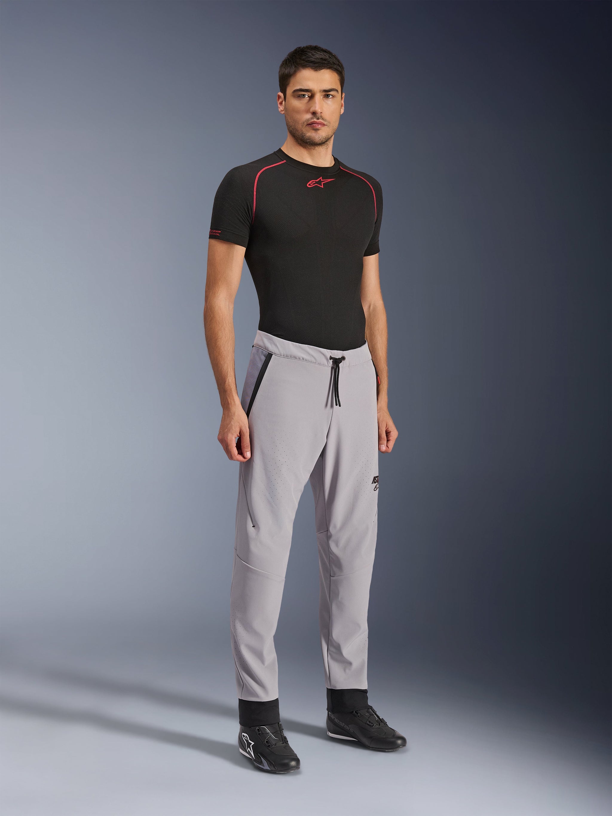 Alpinestars Aeroshell Airflow Broek, motorbroek, Mid Gray, gedragen door een model met een zwart technisch T-shirt en zwarte motorschoenen, voorzien van laser-geperforeerde ventilatiezones, een elastische tailleband met trekkoord en een gestroomlijnde sportieve pasvorm.