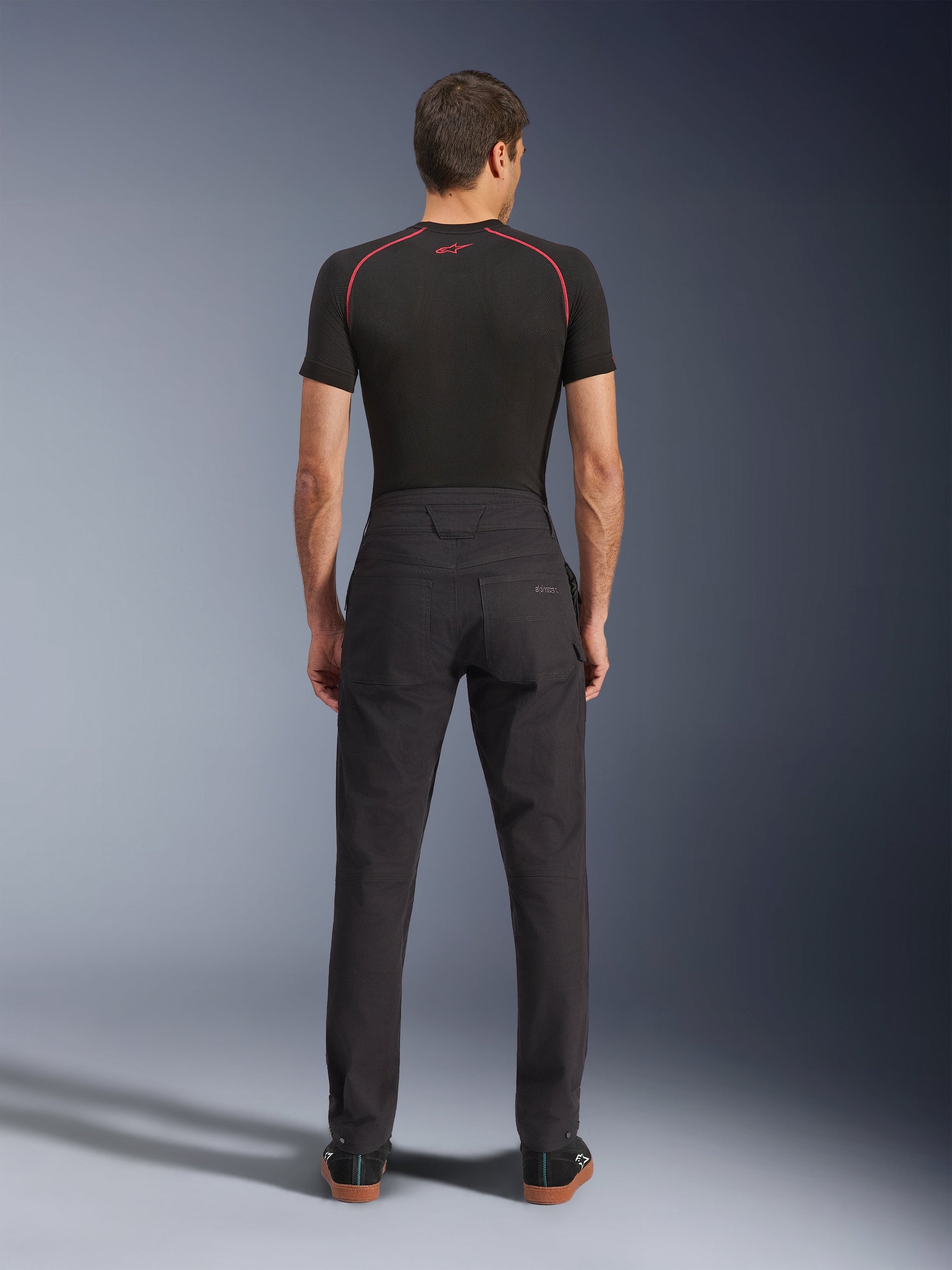 Alpinestars Flex-AST Canvas Pants, urban motorbroek, zwart, achteraanzicht met op werkkleding geïnspireerd ontwerp, achterzakken en verlengde rugstijging, gedragen met een zwarte basislaag en sneakers, ergonomische rijpasvorm
