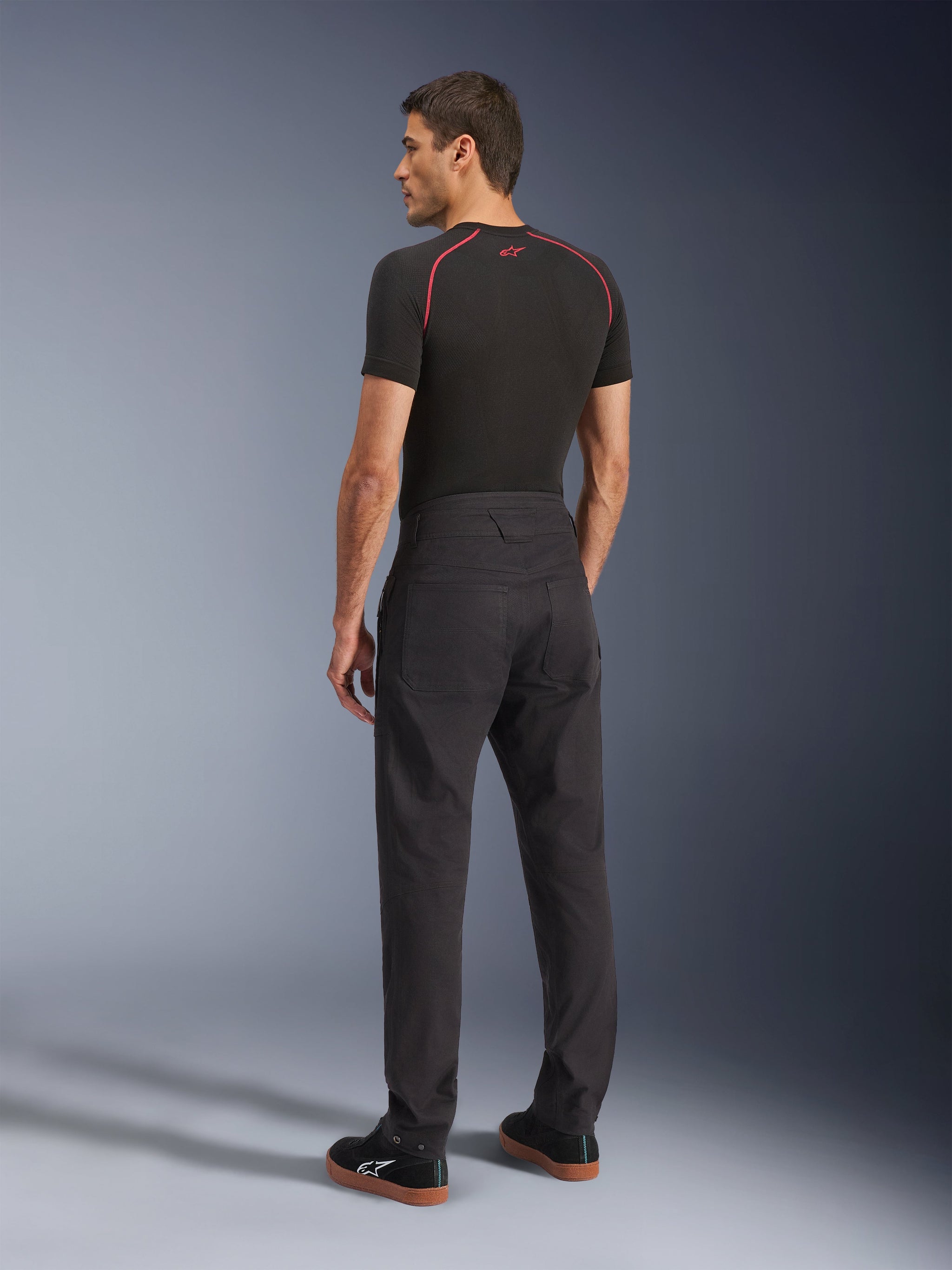 Alpinestars Flex-AST Canvas broek, zwarte urban motorbroek, achteraanzicht op model, gecombineerd met een zwarte basislaag met korte mouwen en zwarte sneakers, voorzien van een op werkkleding geïnspireerd ontwerp met grote achterzakken en een moderne rechte pasvorm.