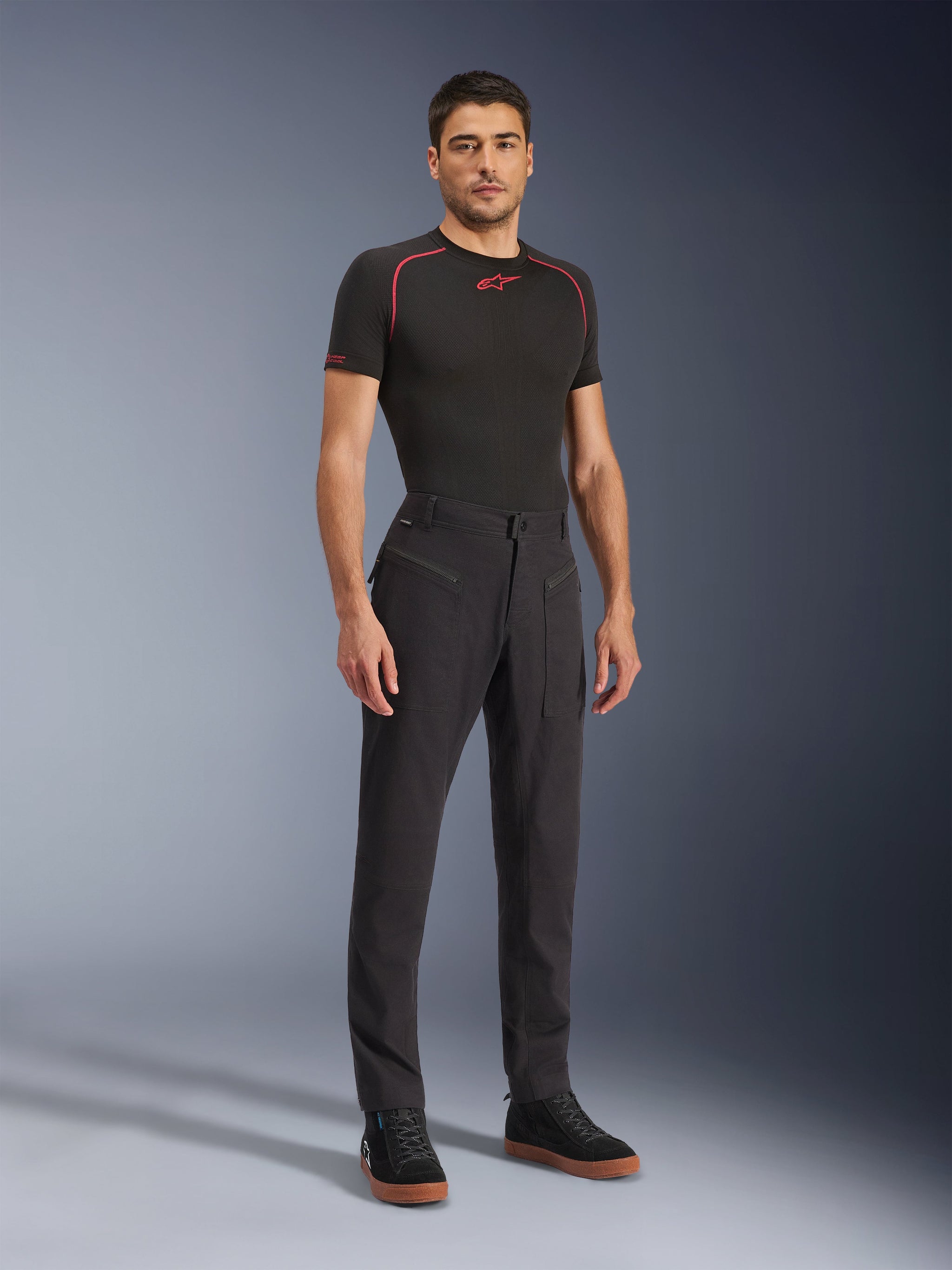 Alpinestars Flex-AST Canvas broek, urbane motorbroek, zwart, gedragen met een zwarte basislaag en casual hoge sneakers, voorzien van grote utility-zakken met rits en een op werkkleding geïnspireerd ontwerp met een ergonomische pasvorm voor het rijden