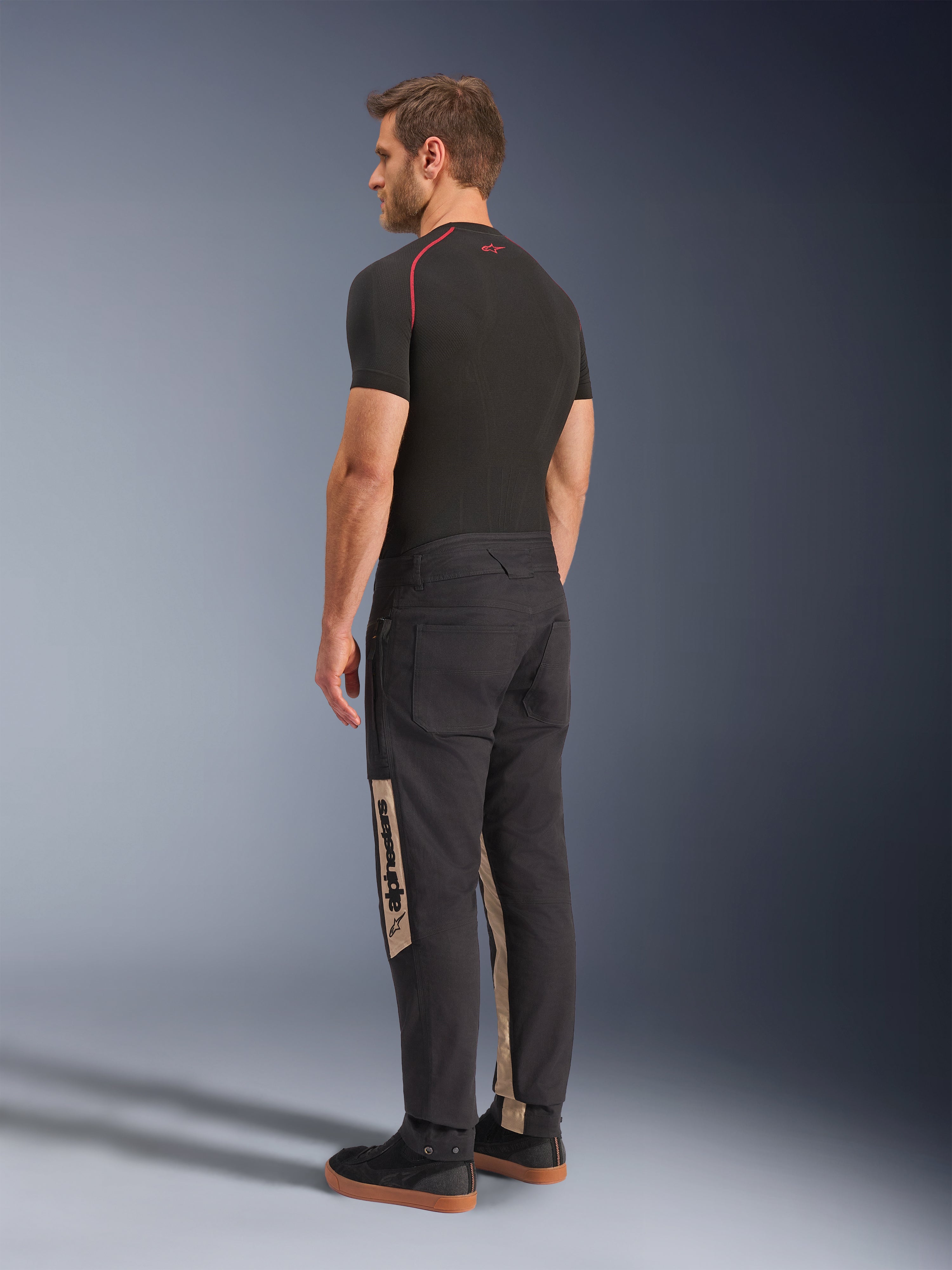 Alpinestars Flex-AST Canvas broek, Black Aluminum, stedelijke motorbroek, met tan zijpanelen met Alpinestars-branding, gedragen met een zwarte basislaag en sneakers, achteraanzicht met op werkkleding geïnspireerde styling en ergonomische motorpasvorm.