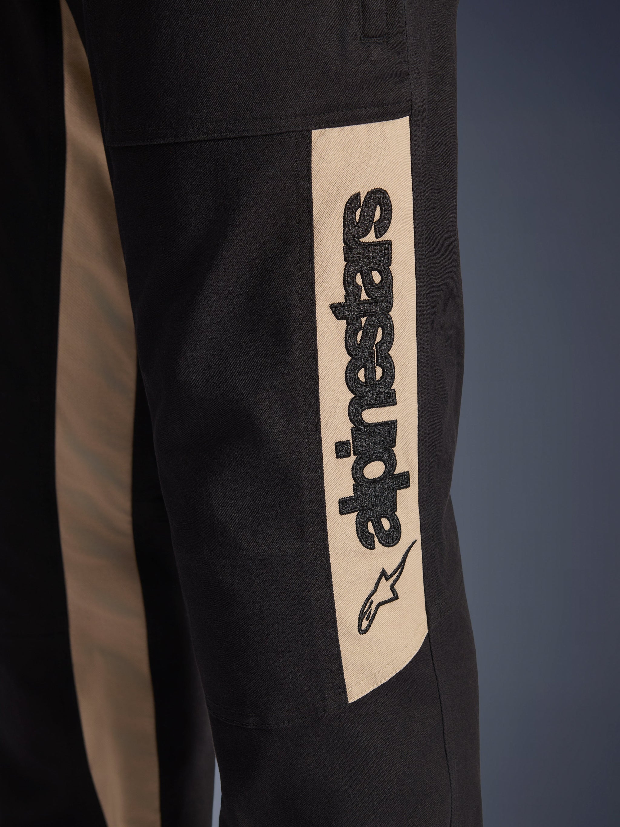 Alpinestars Flex-AST Canvas Pants, close-up detail of the side leg, Black with Aluminum tan accent panel, featuring a vertical black Alpinestars logo and star icon, durable polycotton twill for urban motorcycle riding