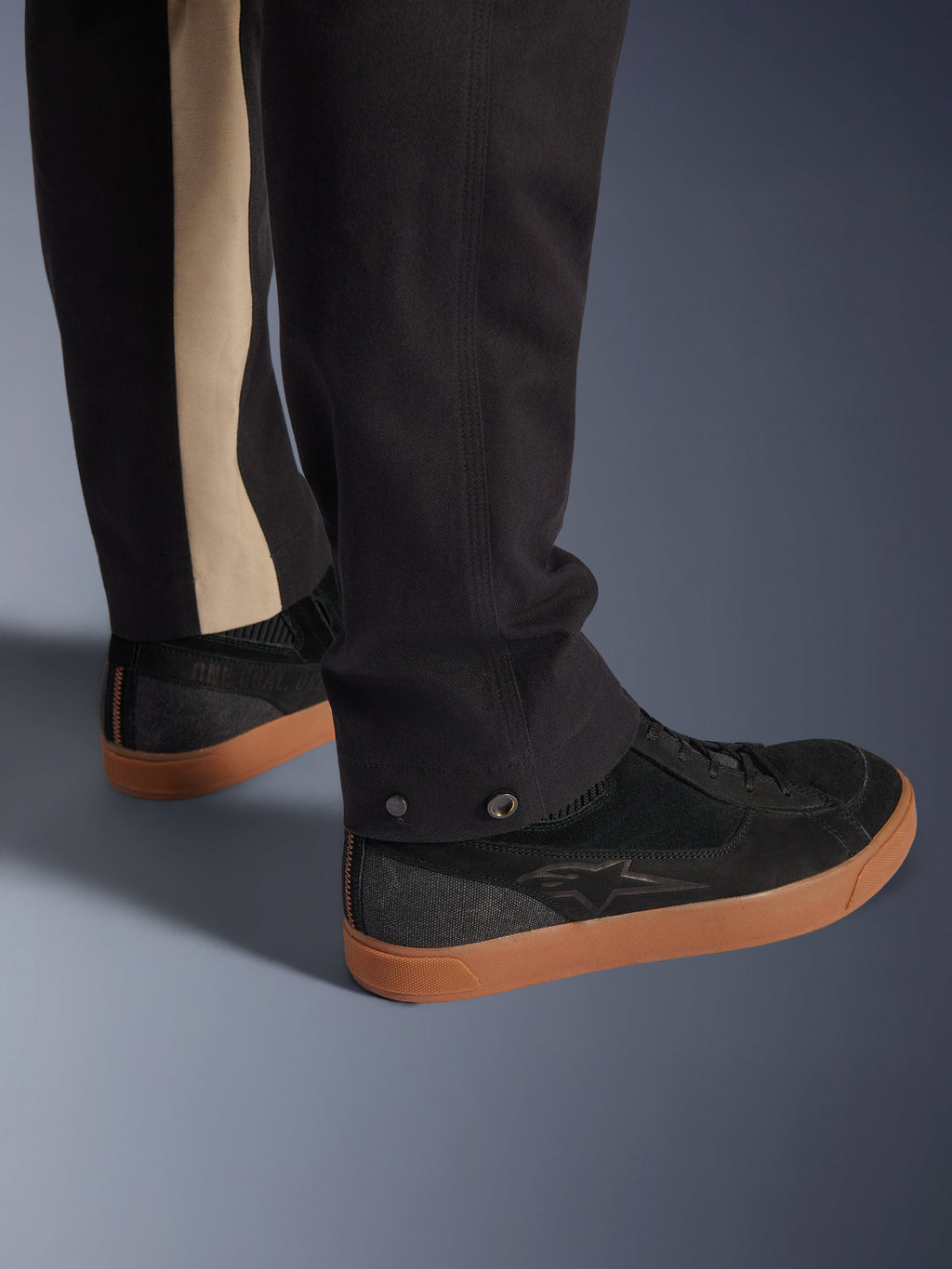 Alpinestars Flex-AST Canvas Broek in Black Aluminum, detailopname van het onderbeen met drukknoopverstellers bij de opening en een verticale aluminiumkleurige streep, gecombineerd met zwarte motorsneakers met gumzolen, stedelijke motorkleding.