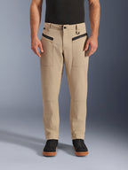 Alpinestars Flex-AST Canvas broek, urban motorbroek, bruin, heritage werkkleding-stijl, voorzien van zwarte horizontale voorzakken met ritssluiting en D-ring aan de riemlus, gedragen met zwart shirt en schoenen, vooraanzicht studio