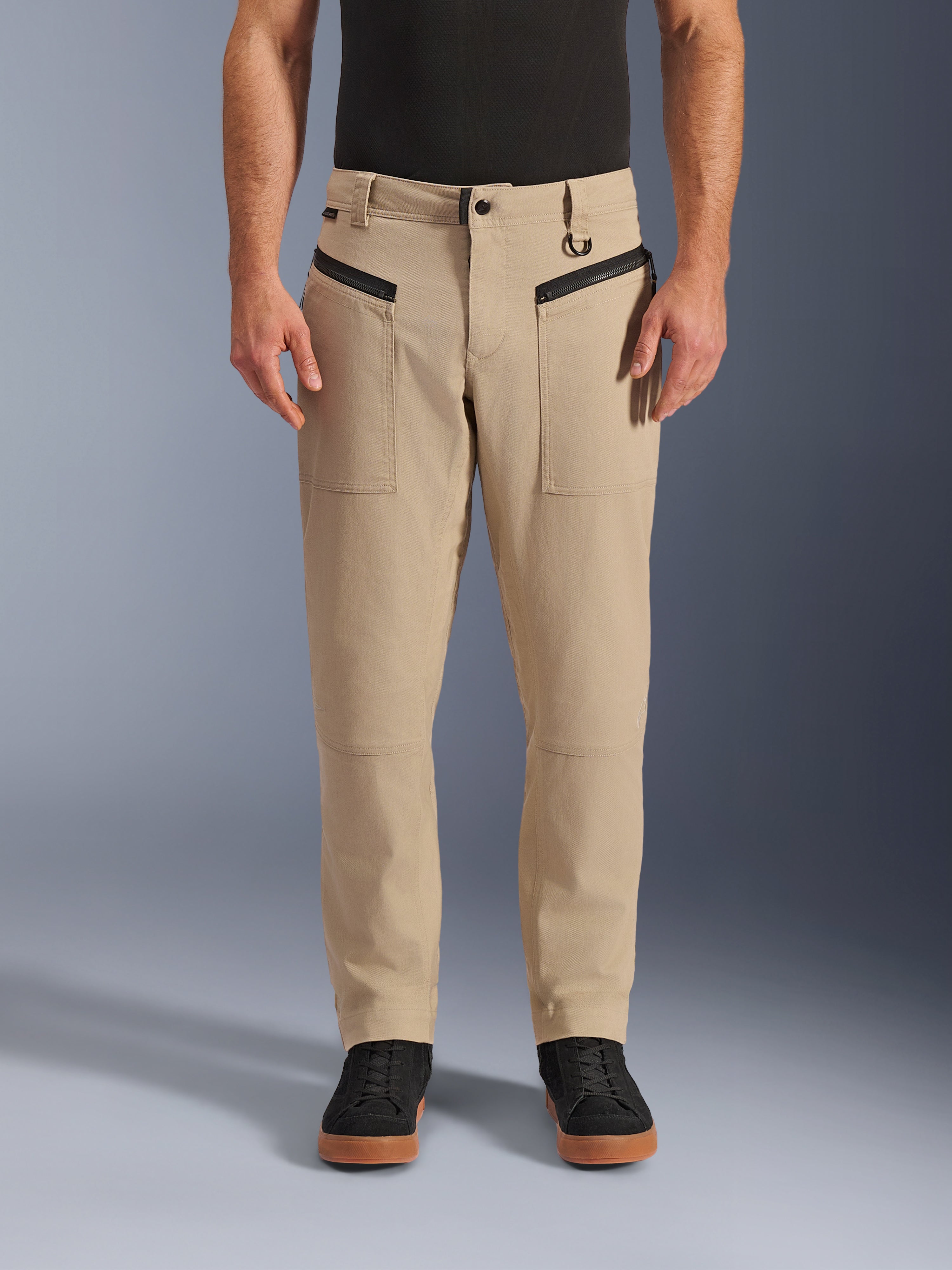 Alpinestars Flex-AST Canvas broek, urban motorbroek, bruin, heritage werkkleding-stijl, voorzien van zwarte horizontale voorzakken met ritssluiting en D-ring aan de riemlus, gedragen met zwart shirt en schoenen, vooraanzicht studio