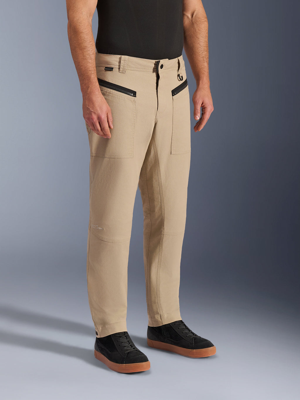 Alpinestars Flex-AST Canvas broek, stedelijke motorbroek, tan bruin, met heritage workwear-styling, voorzakken met ritssluiting en ergonomische kniepanelen, gedragen met zwarte sneakers en een zwart T-shirt, eigentijdse stedelijke motoruitrusting