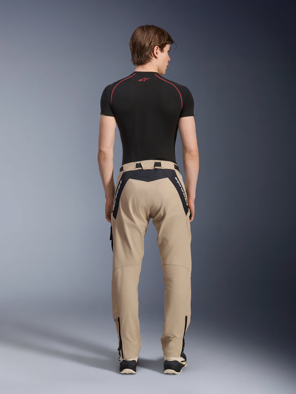 Alpinestars Acteon broek in Sand, moderne urban motorbroek van achteren gezien, voorzien van zwarte panelen met witte Alpinestars-branding, taps toelopende pasvorm met ritsen aan de zoom, gedragen met een zwarte basislaag en sneakers.