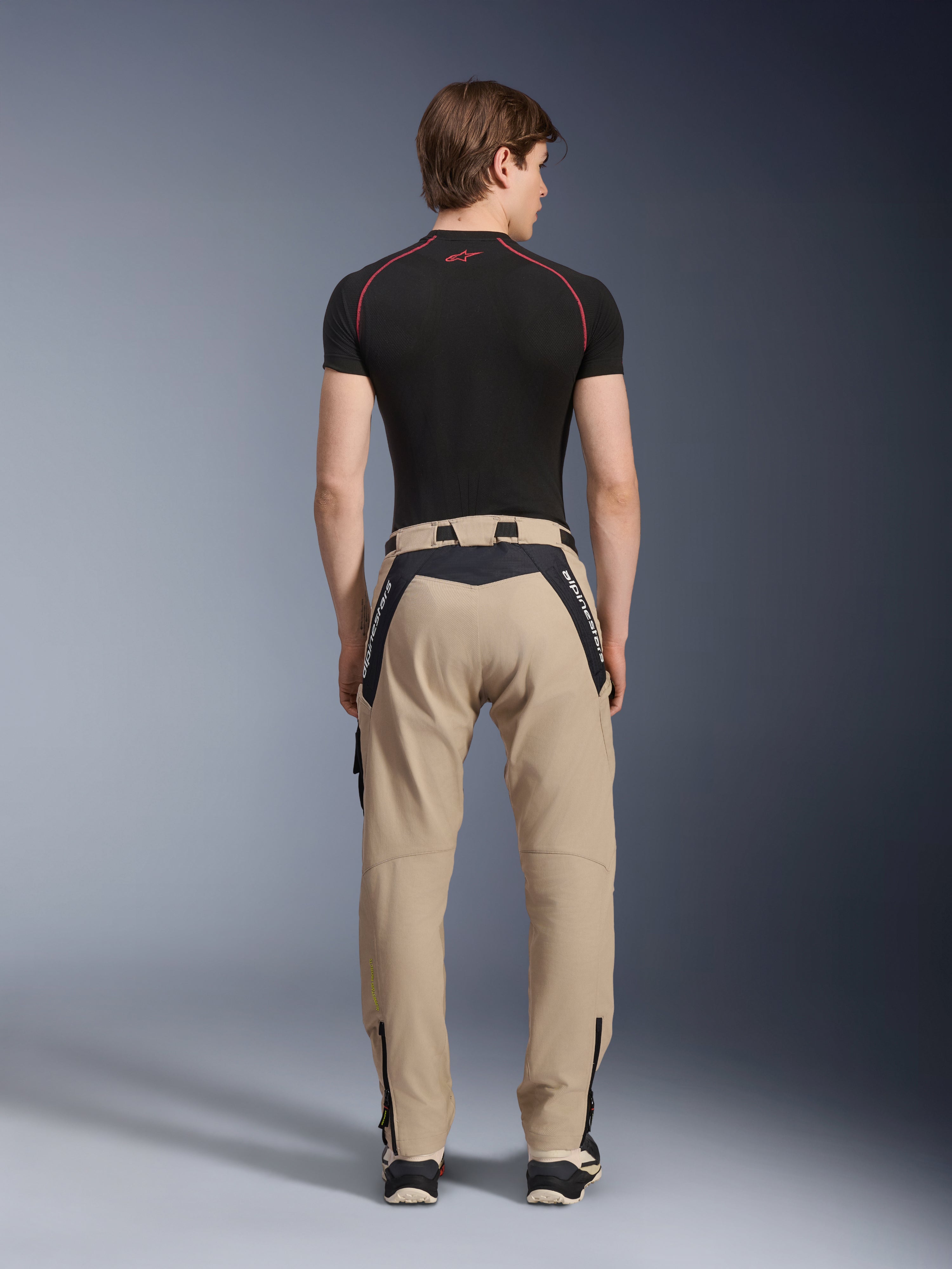 Alpinestars Acteon broek in Sand, moderne urban motorbroek van achteren gezien, voorzien van zwarte panelen met witte Alpinestars-branding, taps toelopende pasvorm met ritsen aan de zoom, gedragen met een zwarte basislaag en sneakers.