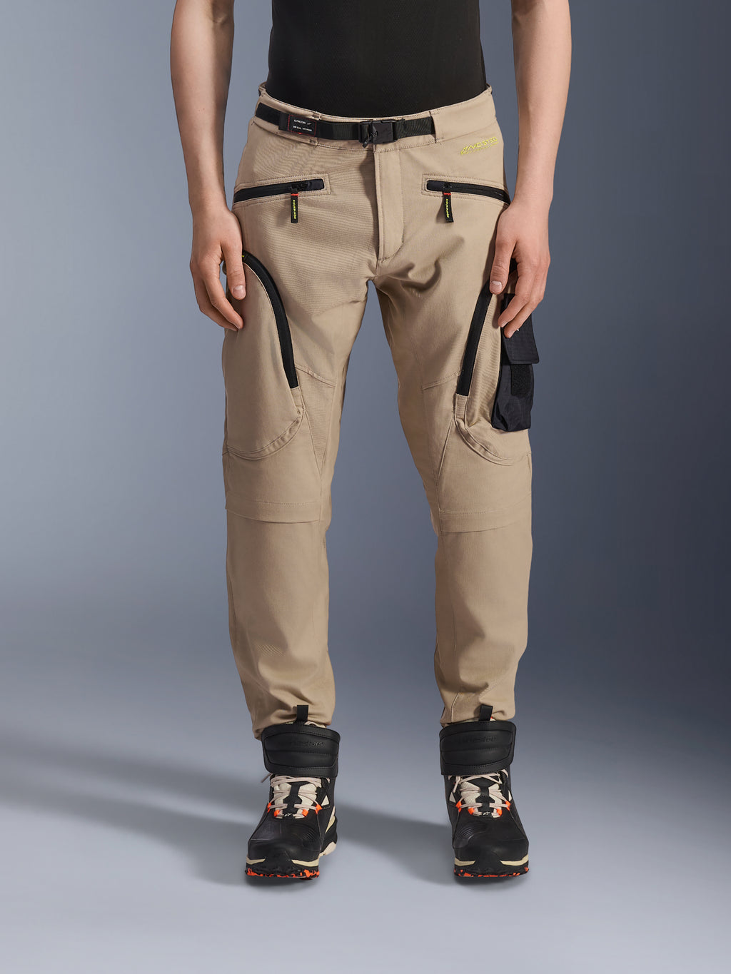 Alpinestars Acteon broek, urban motorrijbroek, zandbeige, met een cargo-ontwerp met meerdere zakken, zwarte omgekeerde spiraalritsen, geïntegreerde riem en een taps toelopende pasvorm, gedragen met zwarte en oranje laarzen.