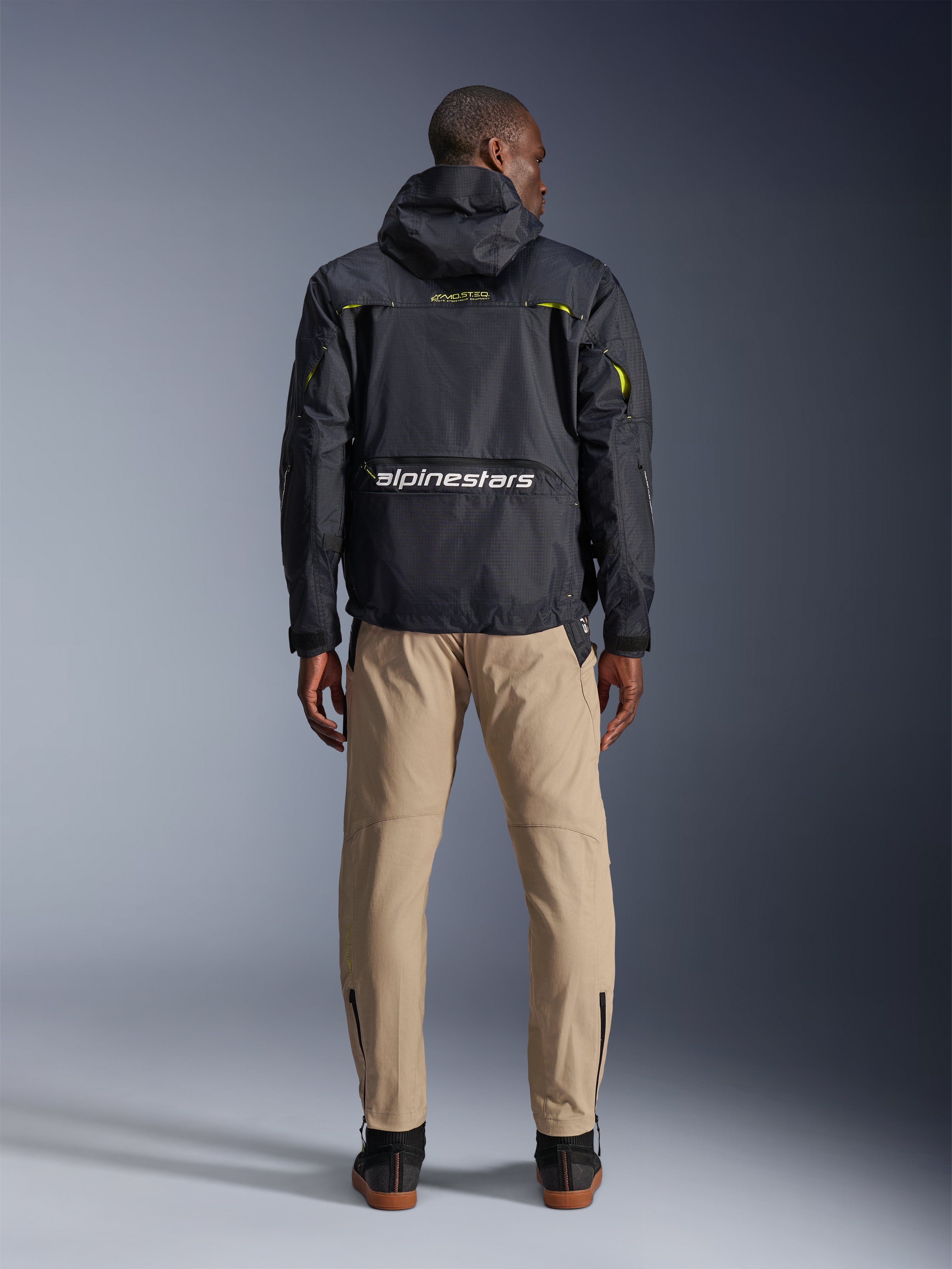 Alpinestars Acteon broek, urban motorbroek, Sand, van achteren afgebeeld op een model dat een zwart technisch jack met capuchon en donkere laarzen draagt, voorzien van taps toelopende broekspijpen met enkelritsen en een strak, modern silhouet.