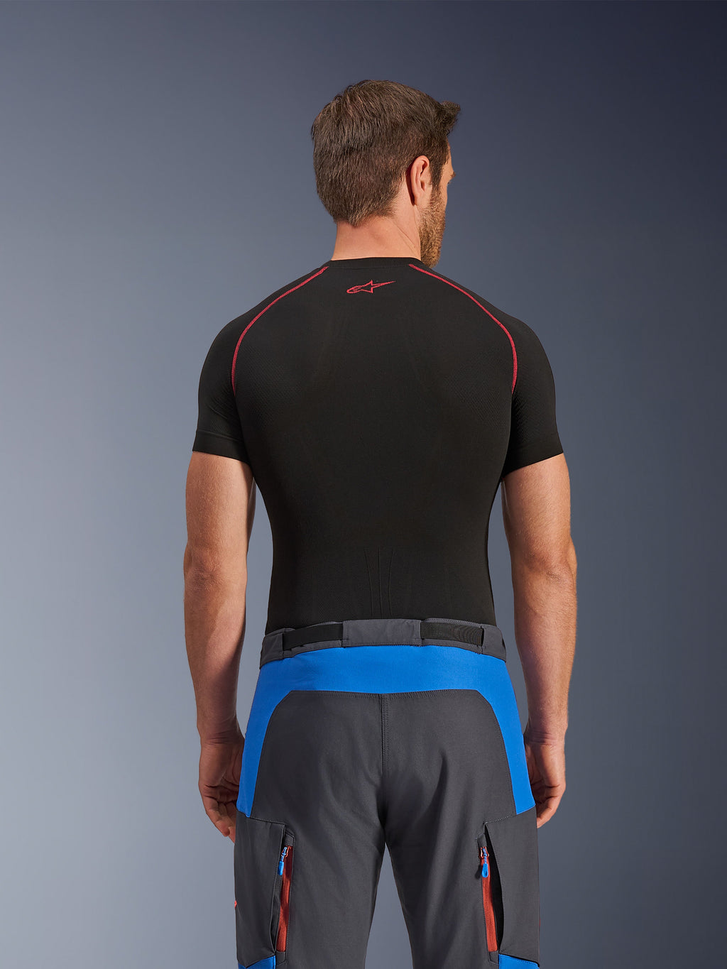 Alpinestars Maxdura Dual broek, adventure motorbroek, koningsblauw en diepgrijs, achteraanzicht, gedragen met een zwarte basislaag met korte mouwen, met blauwe accentpanelen op de bovenrug en oranje cargozakken met rits, technische outdooruitrusting voor dual-sport rijden