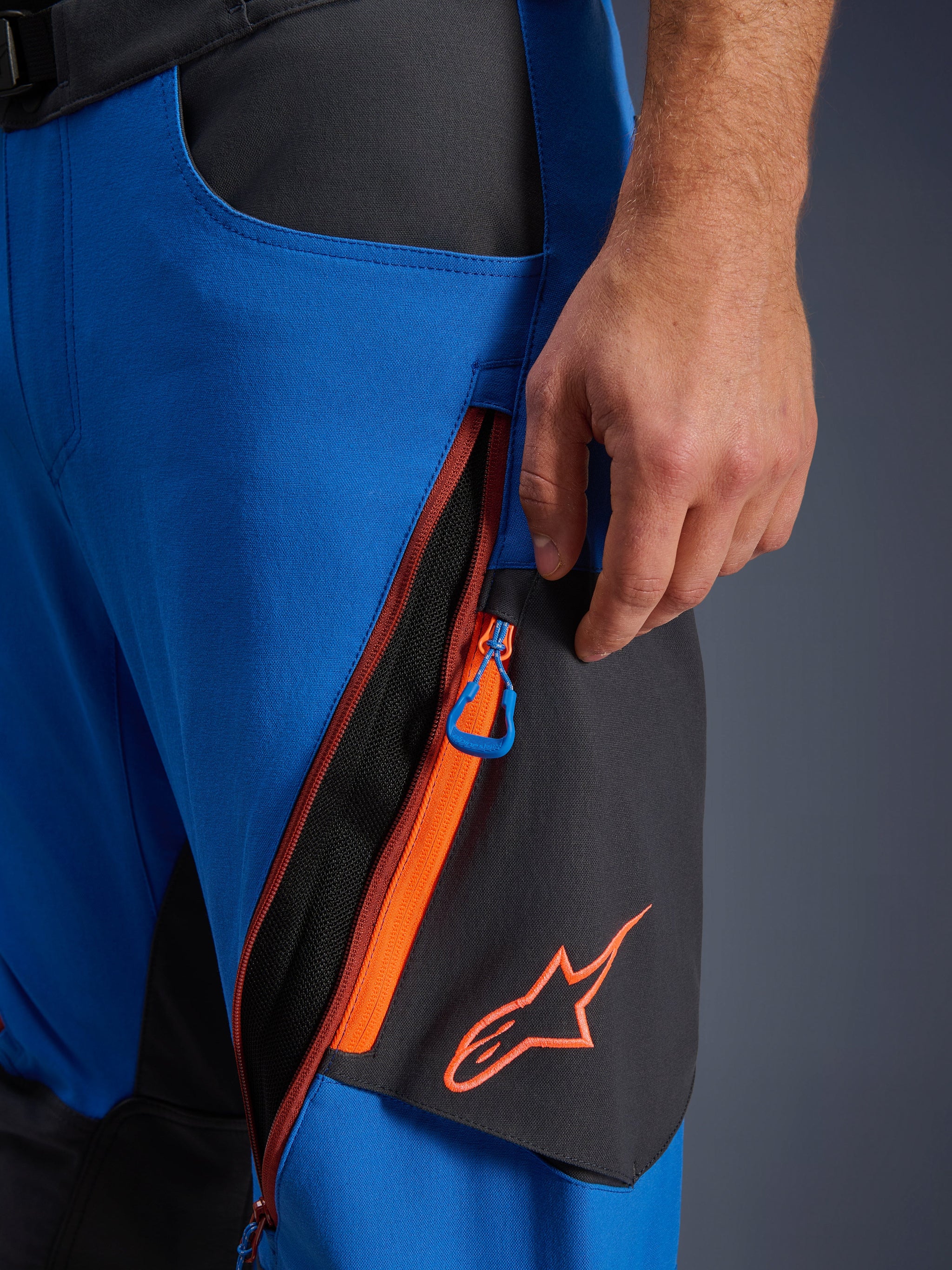 Alpinestars Maxdura Dual Pants detail, adventure motorbroek, Royal Blue en Deep Gray, close-up van ritsbare dijventilatie met oranje accenten en blauwe trekker, oranje Alpinestars-logo op zwart paneel, duurzame stretch polyamide constructie