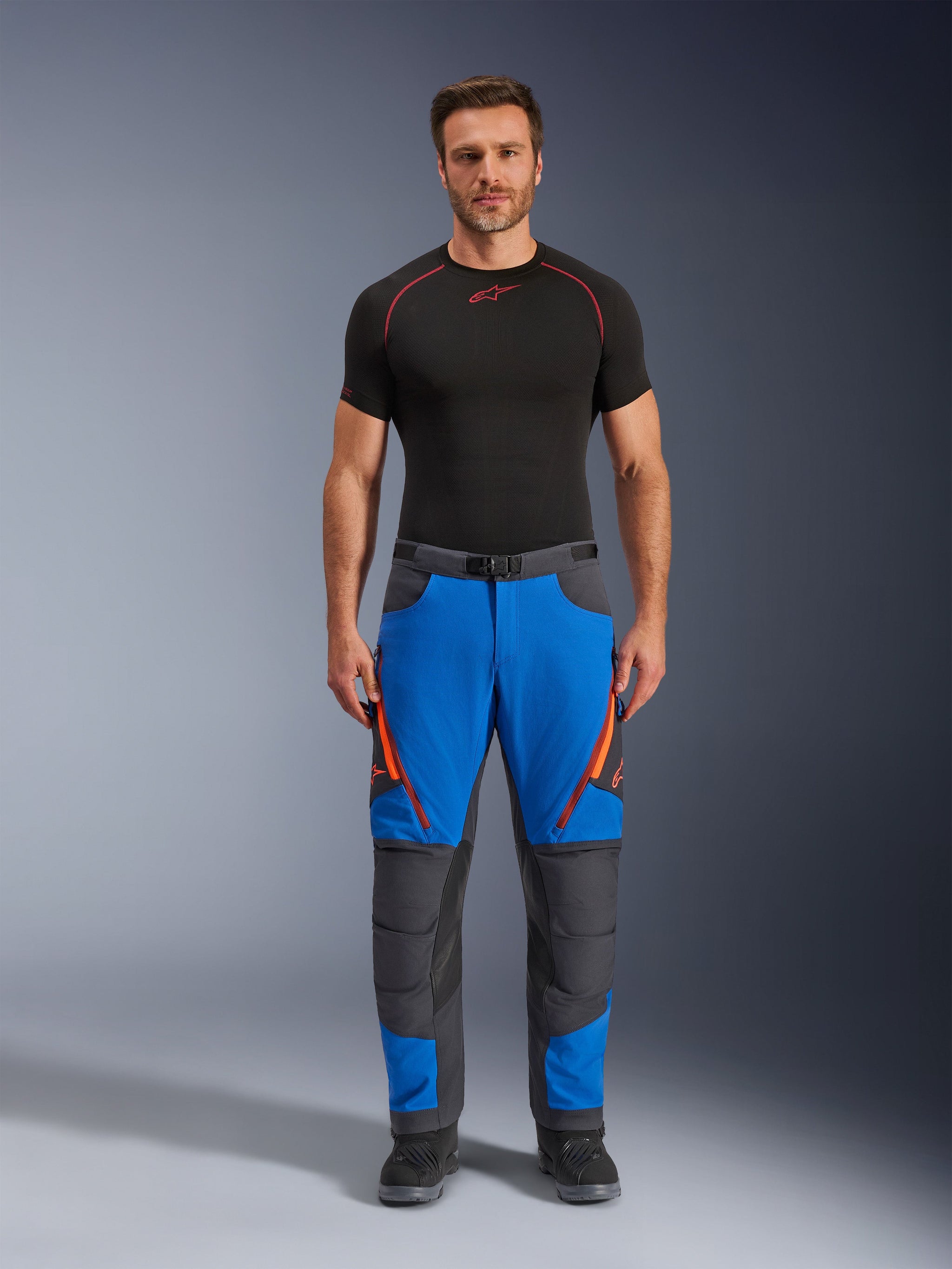 Alpinestars Maxdura Dual Pants, adventure motorbroek, koningsblauw en donkergrijs met oranje ritsaccenten, gedragen met een zwarte Alpinestars basislaag en zwarte laarzen, voorzien van verstevigde kniepanelen en een technisch off-road ontwerp