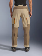 Alpinestars Maxdura Dual broek, Walnut Aluminum, achteraanzicht van adventure motorbroek met beige en aardetinten, ventilatieopeningen met oranje ritsen en over-de-laars ontwerp, gedragen met zwarte Alpinestars laarzen.