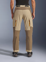 Alpinestars Maxdura Dual broek, Walnut Aluminum, achteraanzicht van adventure motorbroek met beige en aardetinten, ventilatieopeningen met oranje ritsen en over-de-laars ontwerp, gedragen met zwarte Alpinestars laarzen.