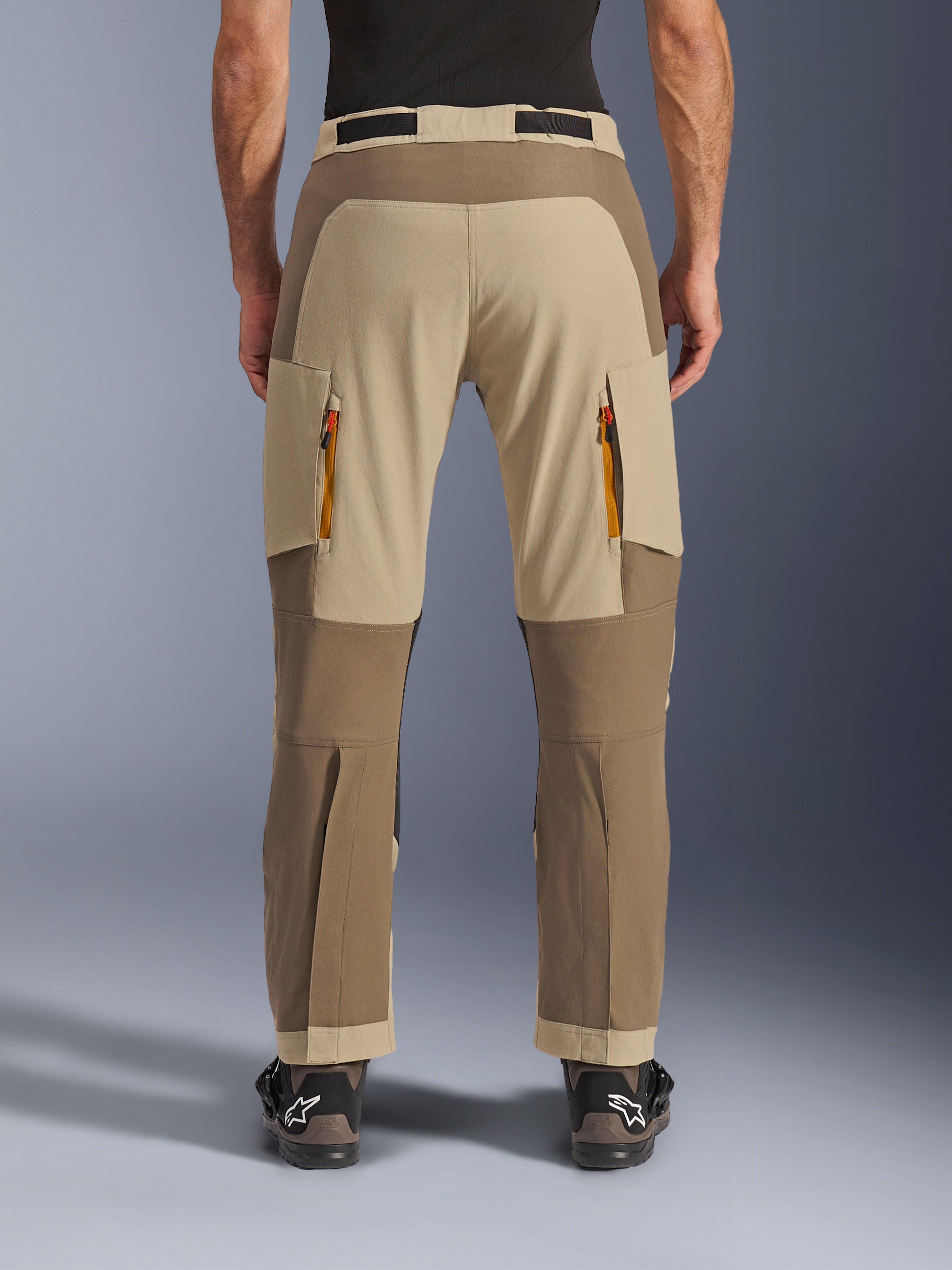 Alpinestars Maxdura Dual broek, Walnut Aluminum, achteraanzicht van adventure motorbroek met beige en aardetinten, ventilatieopeningen met oranje ritsen en over-de-laars ontwerp, gedragen met zwarte Alpinestars laarzen.