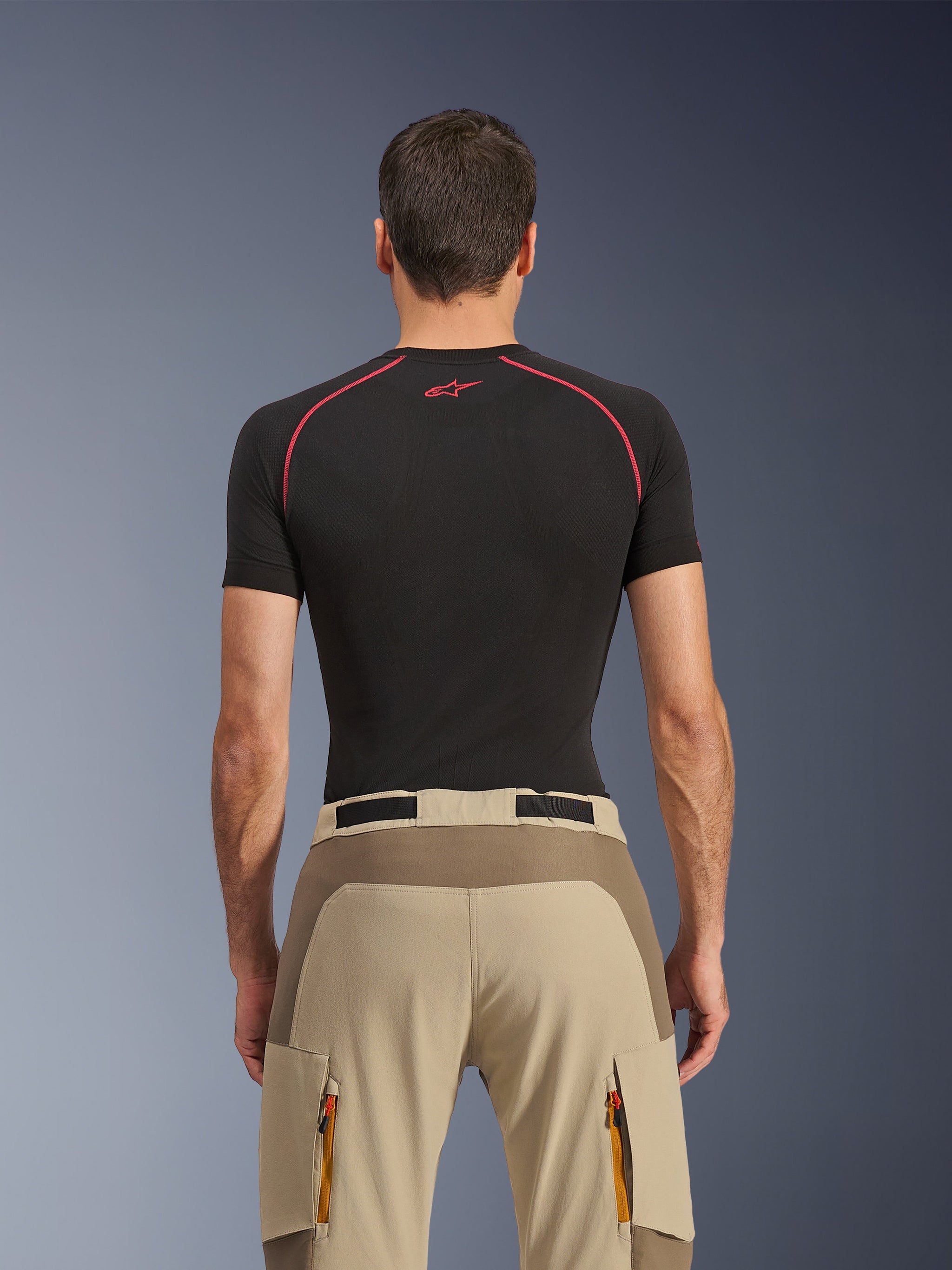 Alpinestars Maxdura Dual broek, adventure motorbroek, Walnut Aluminum, achteraanzicht met verstevigd zitvlakpaneel en beige stretchstof, cargozakken met ritssluiting en oranje trekkers, gedragen met een zwart Alpinestars base layer shirt.