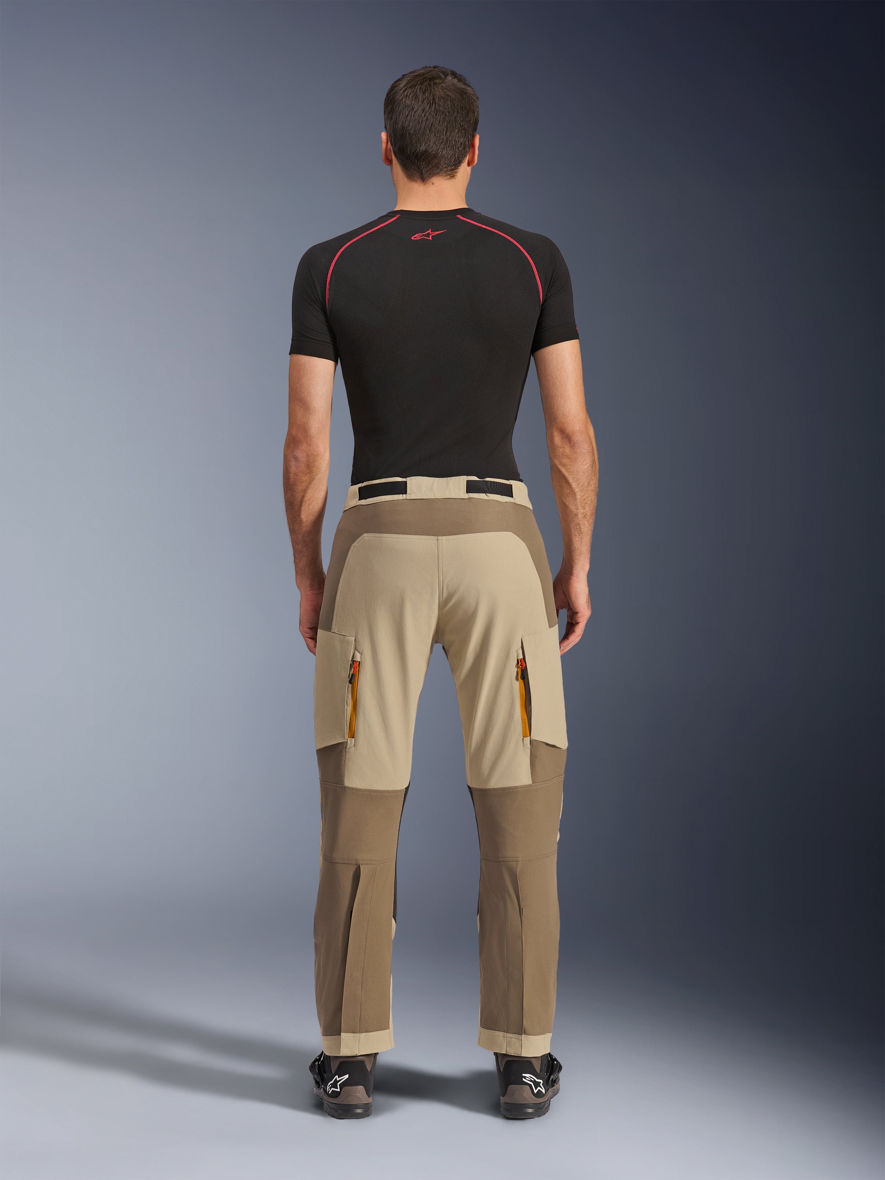 Alpinestars Maxdura Dual Pants in Walnut Aluminum, achteraanzicht met tweekleurig bruin en beige ontwerp, adventure motorbroek met cargozakken met ritssluiting en versterkt zitvlak, gedragen met een zwart compressieshirt en motorlaarzen