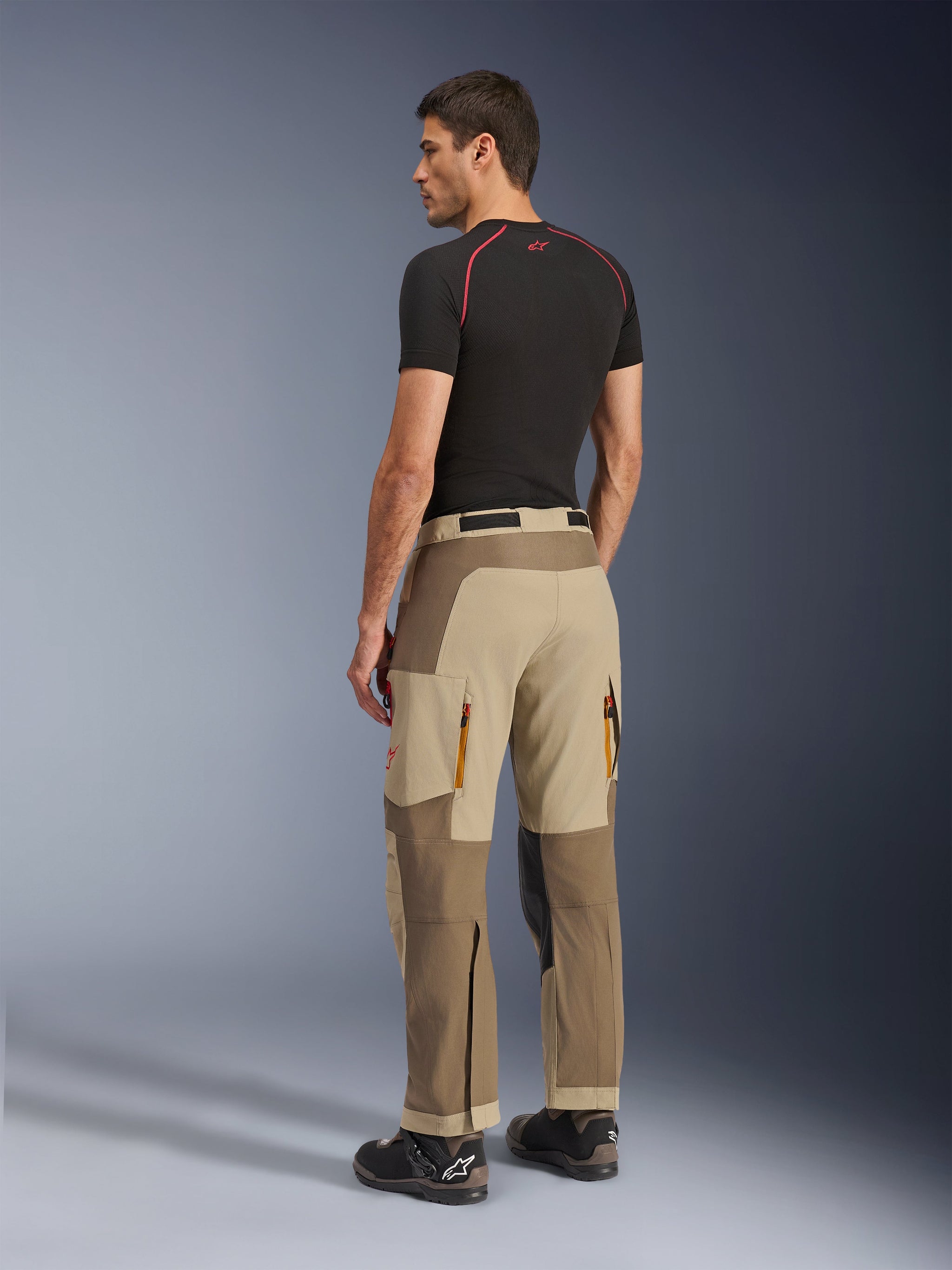 Alpinestars Maxdura Dual Pants, adventure- en dual-sport motorbroek, Walnut Aluminum, achteraanzicht gedragen door een model met een zwarte basislaag en motorlaarzen, voorzien van een multi-paneel constructie met ventilatieopeningen met rits aan de achterzijde en versterkte overlays.