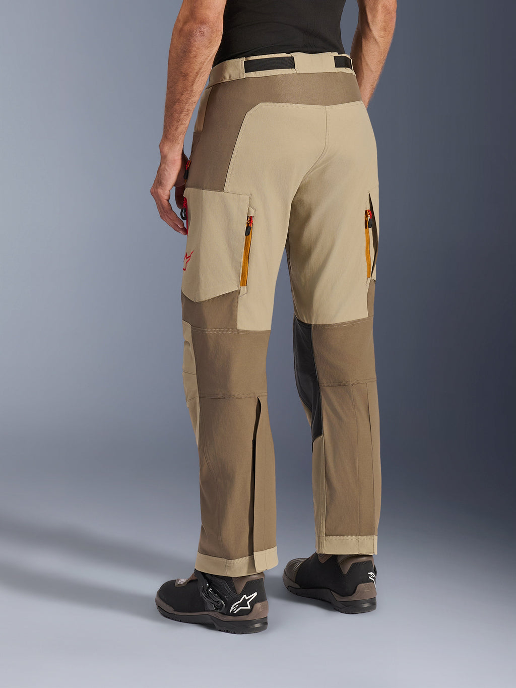Alpinestars Maxdura Dual Pants in Walnut Aluminum, achteraanzicht, adventure motorbroek met beige en bruine panelen, ventilatieopeningen met rits aan de achterzijde, grote cargozakken en een over-de-laars ontwerp gedragen met zwarte en bruine laarzen