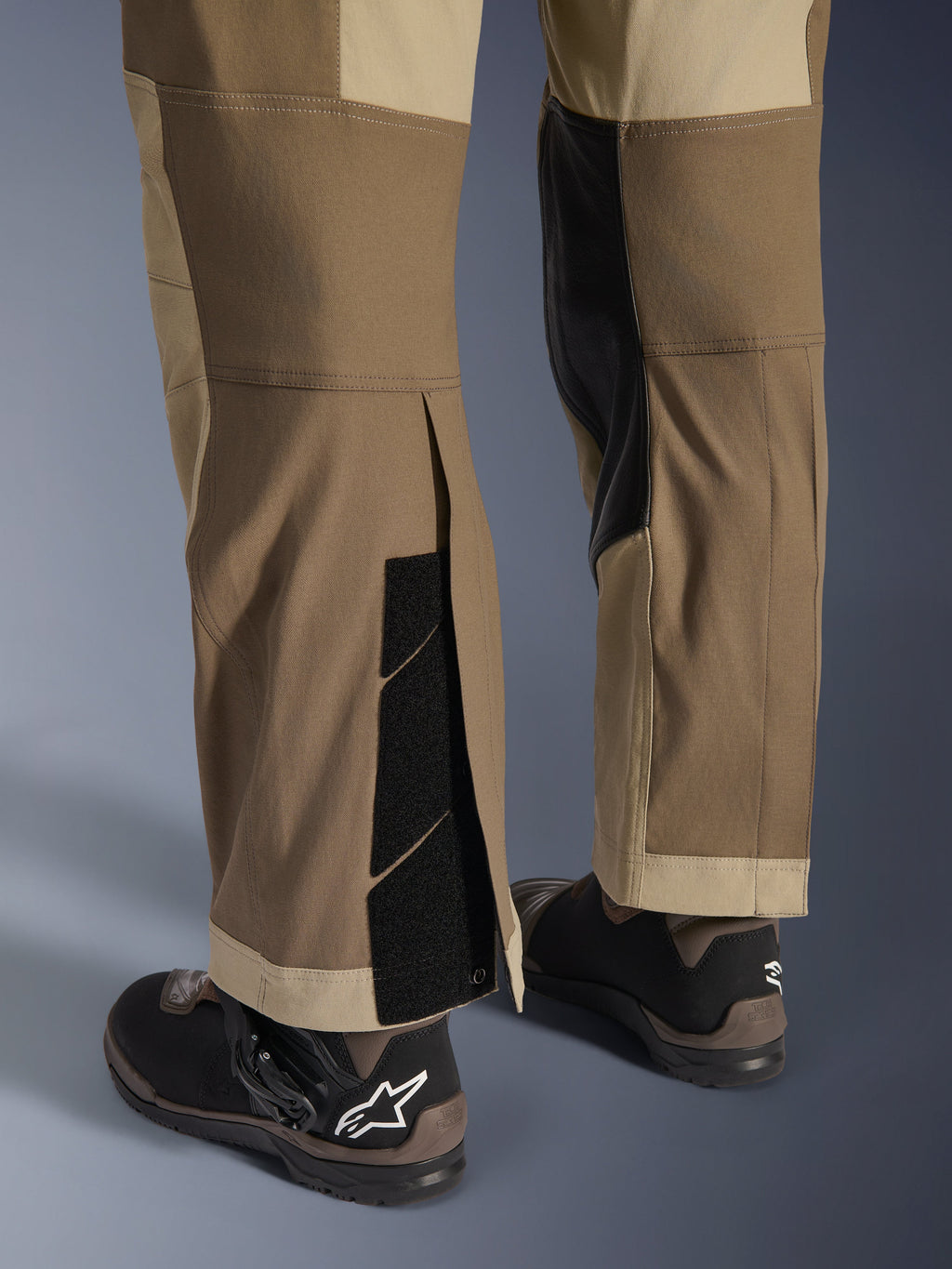 Alpinestars Maxdura Dual broek in Walnut Aluminum, detailweergave van het onderbeen en de zoom met een over-de-laars ontwerp, verstelbare klittenbandsluitingen en verstevigde binnenpanelen, gecombineerd met zwarte motorlaarzen.