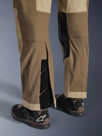 Alpinestars Maxdura Dual broek in Walnut Aluminum, detailweergave van het onderbeen en de zoom met een over-de-laars ontwerp, verstelbare klittenbandsluitingen en verstevigde binnenpanelen, gecombineerd met zwarte motorlaarzen.