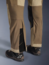 Alpinestars Maxdura Dual broek in Walnut Aluminum, detailweergave van het onderbeen en de zoom met een over-de-laars ontwerp, verstelbare klittenbandsluitingen en verstevigde binnenpanelen, gecombineerd met zwarte motorlaarzen.