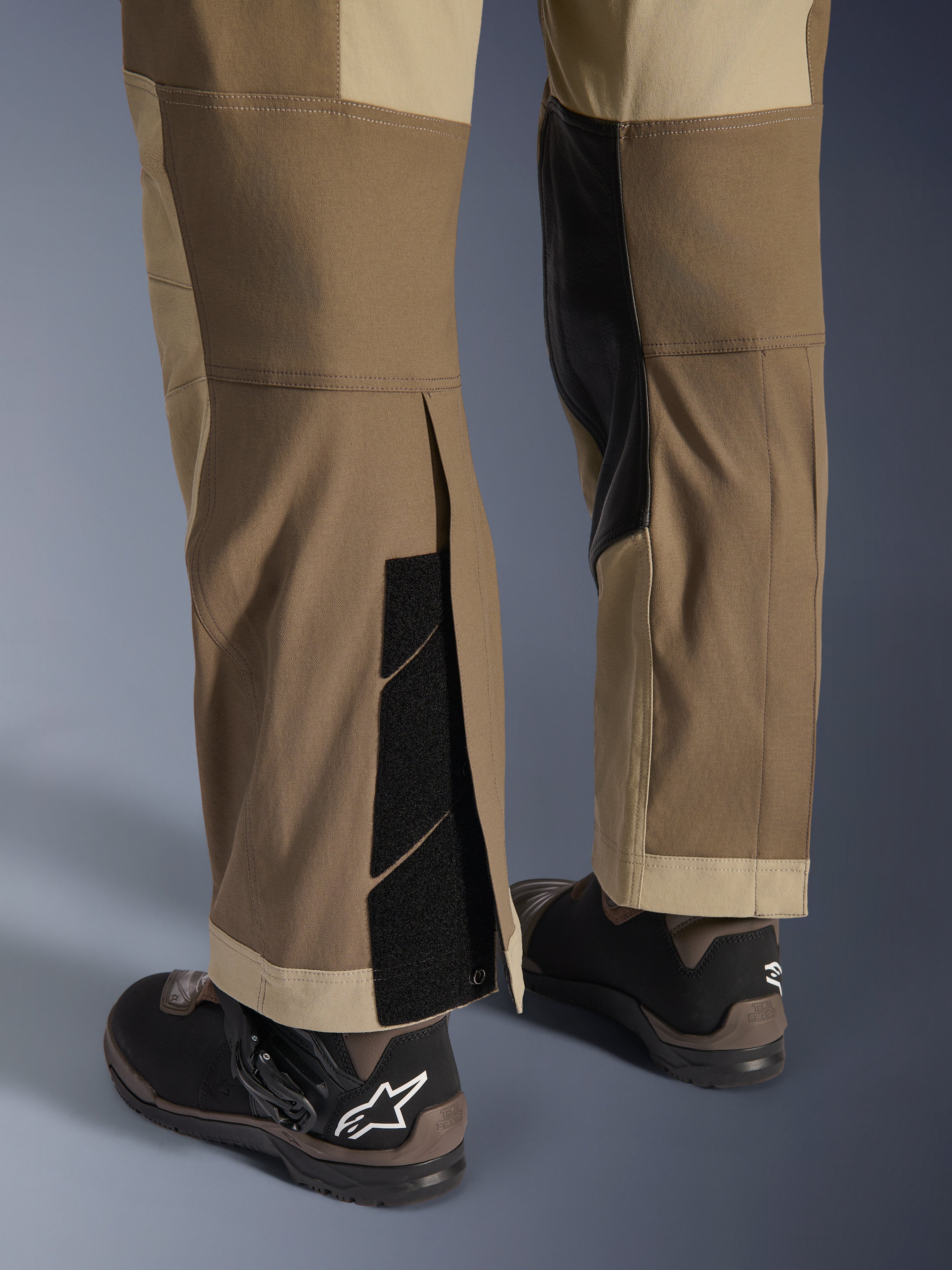 Alpinestars Maxdura Dual broek in Walnut Aluminum, detailweergave van het onderbeen en de zoom met een over-de-laars ontwerp, verstelbare klittenbandsluitingen en verstevigde binnenpanelen, gecombineerd met zwarte motorlaarzen.