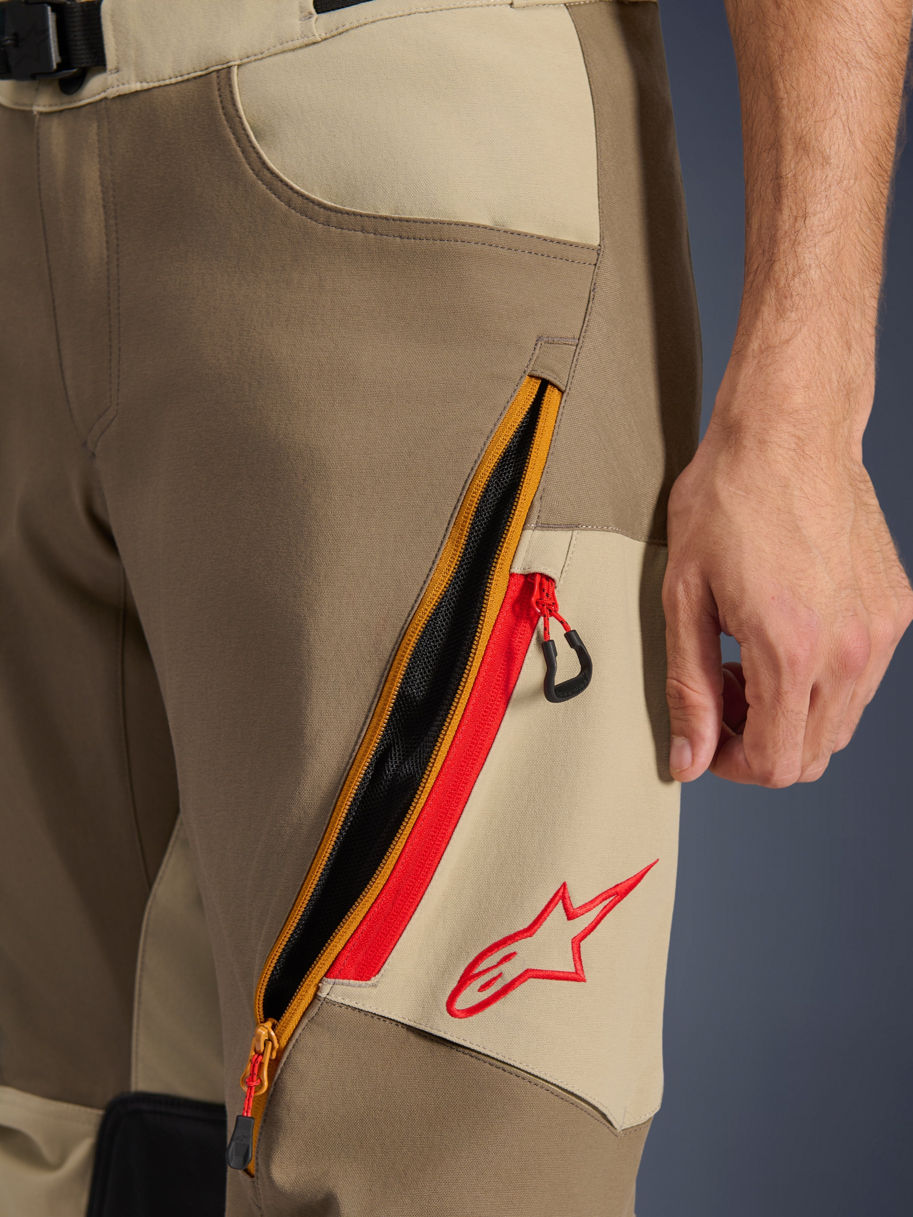 Alpinestars Maxdura Dual broek, Walnut en Aluminum, detailopname van de dij met een groot diagonaal ventilatiesysteem met ritssluiting, rode accenten en een rood Alpinestars-logo op een beige paneel, adventure motorkleding