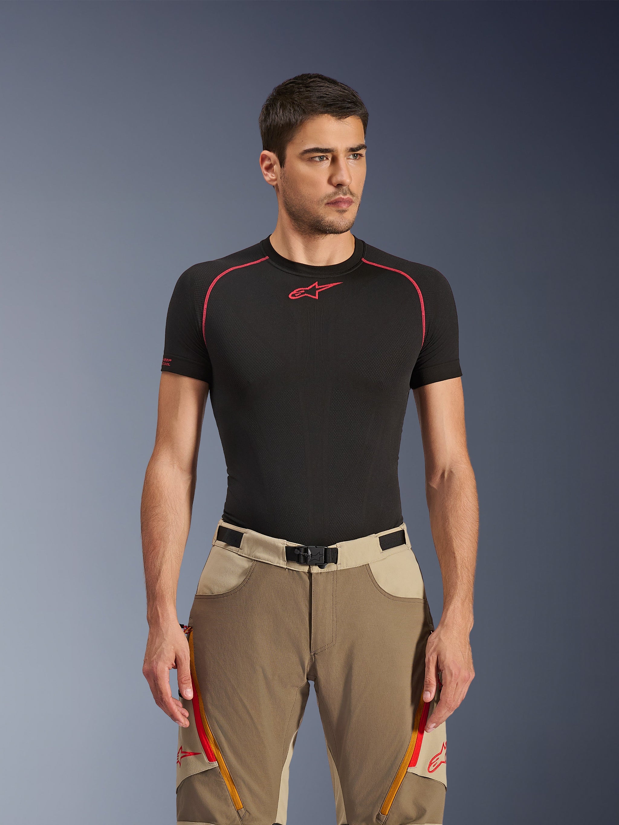 Alpinestars Maxdura Dual Pants in Walnut Aluminum, bruine en beige adventure motorbroek gedragen door een model met een zwart ondershirt, voorzien van oranje dijventilatie met rits, rode logo's en een magnetische gespsluiting, studioportret.