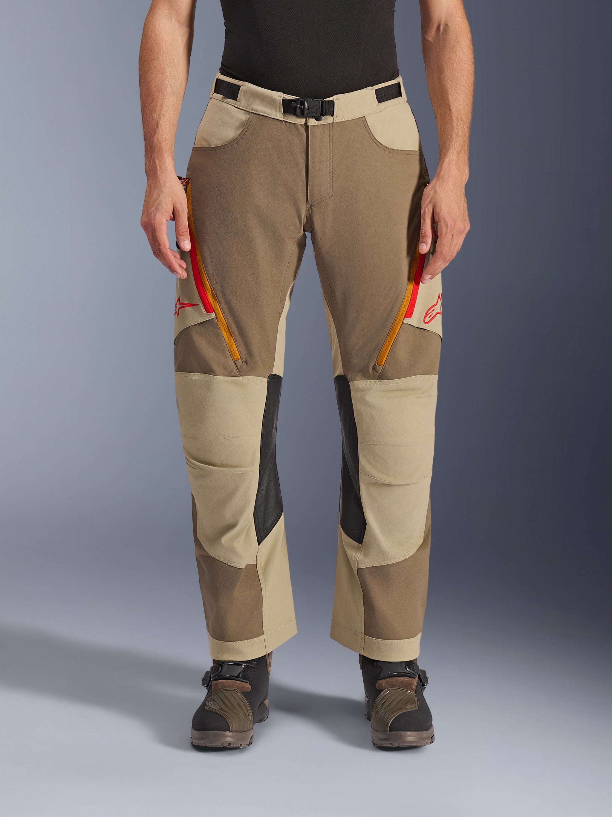 Alpinestars Maxdura Dual Pants, Walnut Aluminum, adventure motorbroek met rood en oranje ventilatieritsen op de dijen, zwarte leren binnenkniepanelen, gedragen met een zwarte basislaag en robuuste laarzen, duurzaam meerkleurig ontwerp voor dual-sport ritten