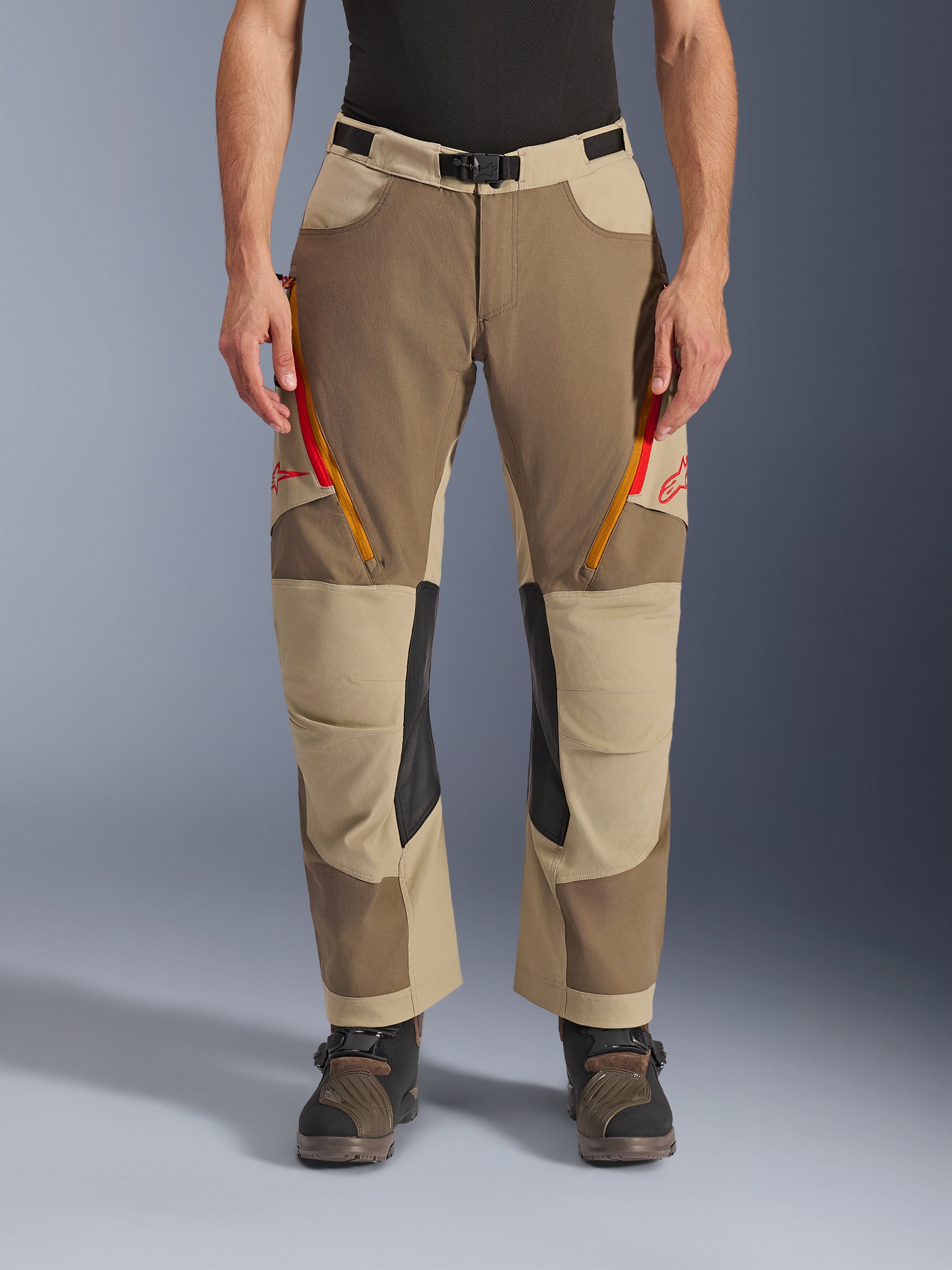 Alpinestars Maxdura Dual Pants, Walnut Aluminum, adventure motorbroek met rood en oranje ventilatieritsen op de dijen, zwarte leren binnenkniepanelen, gedragen met een zwarte basislaag en robuuste laarzen, duurzaam meerkleurig ontwerp voor dual-sport ritten