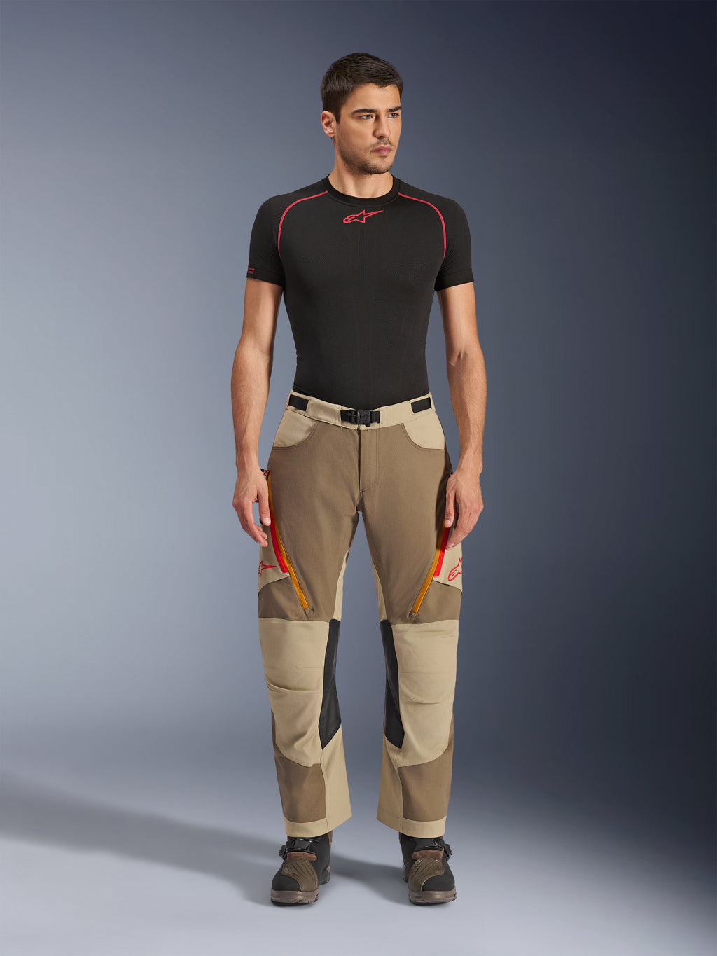 Alpinestars Maxdura Dual broek, adventure- en dual-sport motorbroek, Walnut Aluminum, gedragen met een zwart compressieshirt met korte mouwen en adventure-laarzen, voorzien van oranje ventilatieopeningen met rits en rode logo's op de dijbenen, zwarte tailleriem met gesp, over-de-laars ontwerp voor motorrijden.