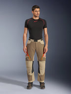 Alpinestars Maxdura Dual broek, adventure- en dual-sport motorbroek, Walnut Aluminum, gedragen met een zwart compressieshirt met korte mouwen en adventure-laarzen, voorzien van oranje ventilatieopeningen met rits en rode logo's op de dijbenen, zwarte tailleriem met gesp, over-de-laars ontwerp voor motorrijden.