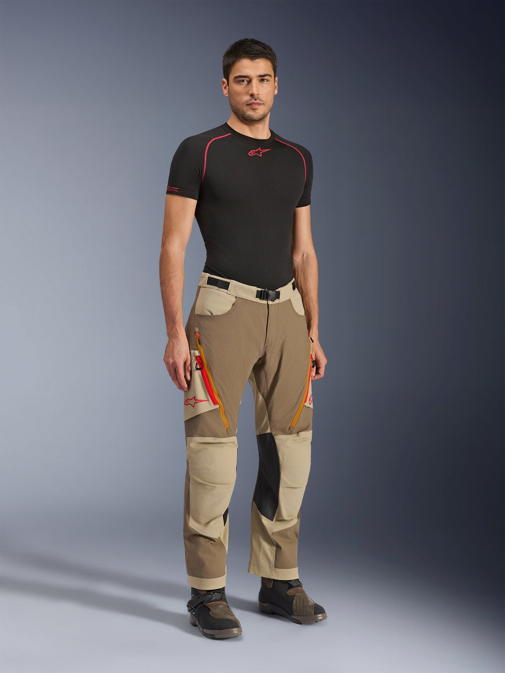 Alpinestars Maxdura Dual Pants in Walnut Aluminum, bruine en beige adventure motorbroek met oranje ritsen en zwarte leren kniepanelen, gedragen met een zwart base-layer T-shirt en adventure-laarzen, vooraanzicht van het volledige lichaam