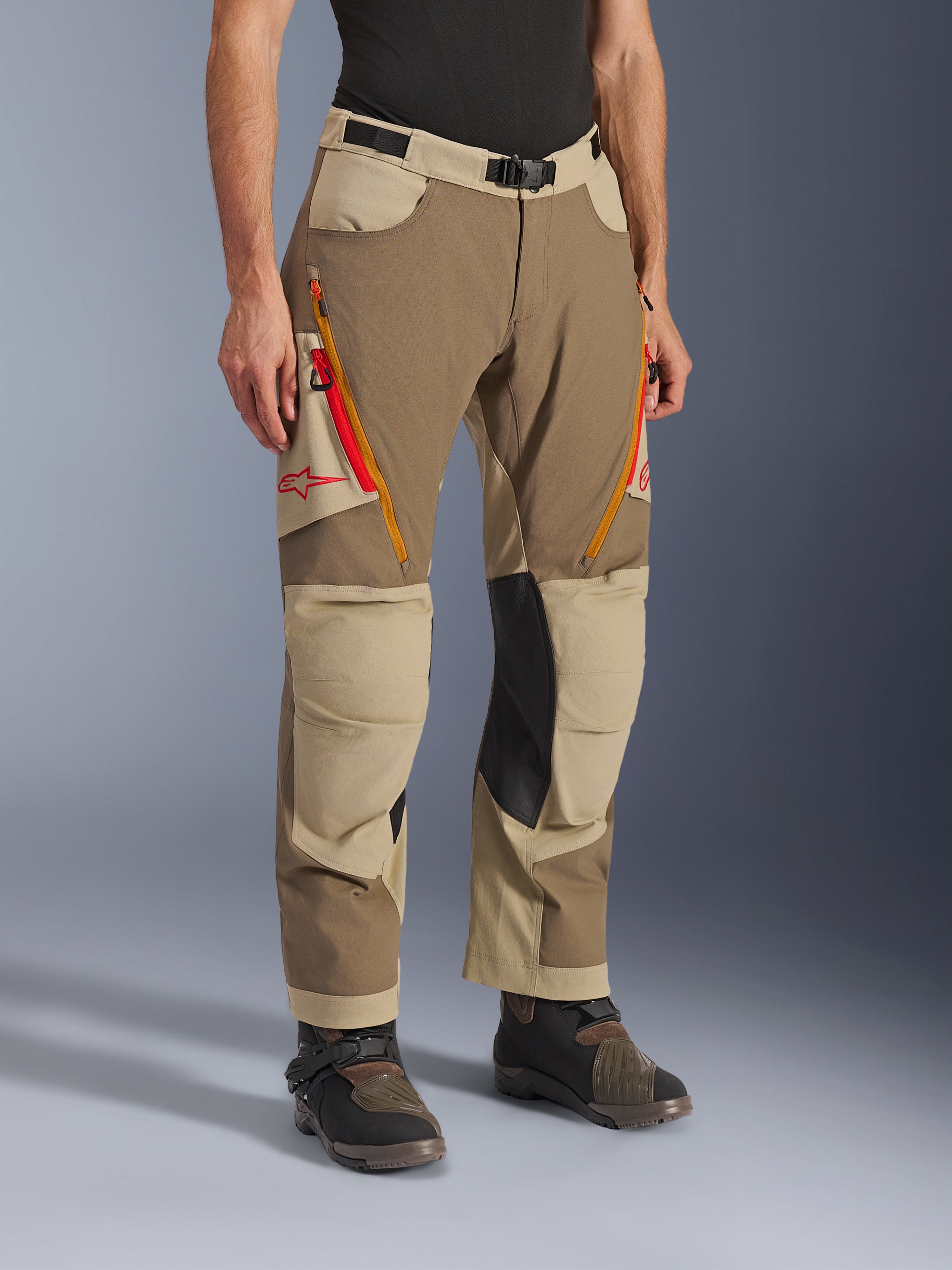 Alpinestars Maxdura Dual Pants, Walnut Aluminum bruine en khaki adventure motorbroek, met oranje en rode ventilatieritsen op de dijen, verstevigde zwarte leren binnenkniepanelen en rood Astars-logo, gedragen met technische laarzen.