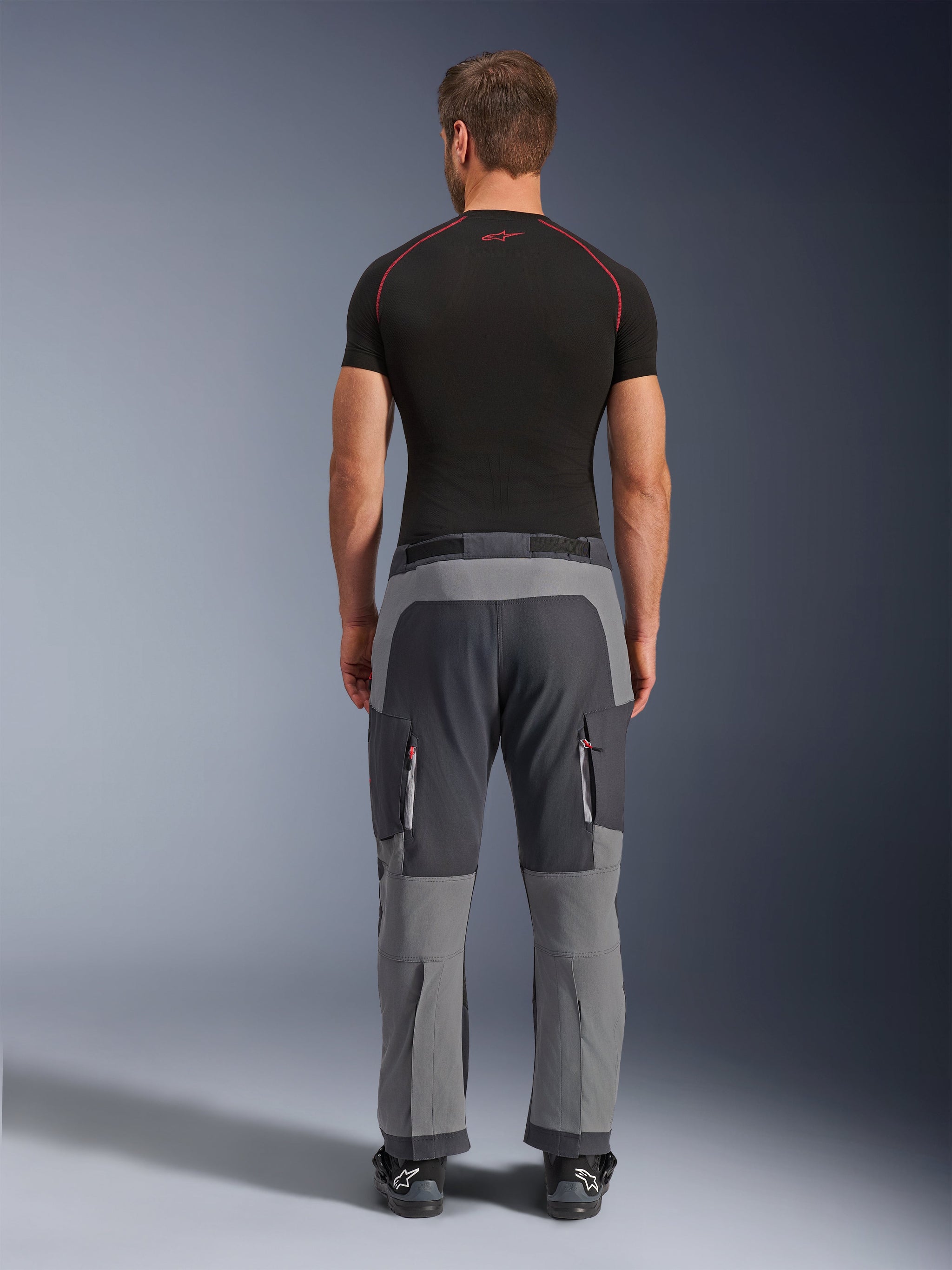Alpinestars Maxdura Dual Pants, donkergrijze en diepgrijze adventure motorbroek, achteraanzicht op model met zwart T-shirt en laarzen, voorzien van meerkleurige grijze panelen en cargozakken met rits en rode accenten.
