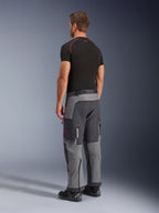 Alpinestars Maxdura Dual Pants, adventure motorbroek, donkergrijs en diepgrijs, gedragen door een model van achteren opzij gezien, met rode accenten op ritsen en logo, versterkte grijze panelen, cargozakken en over-de-laars pasvorm