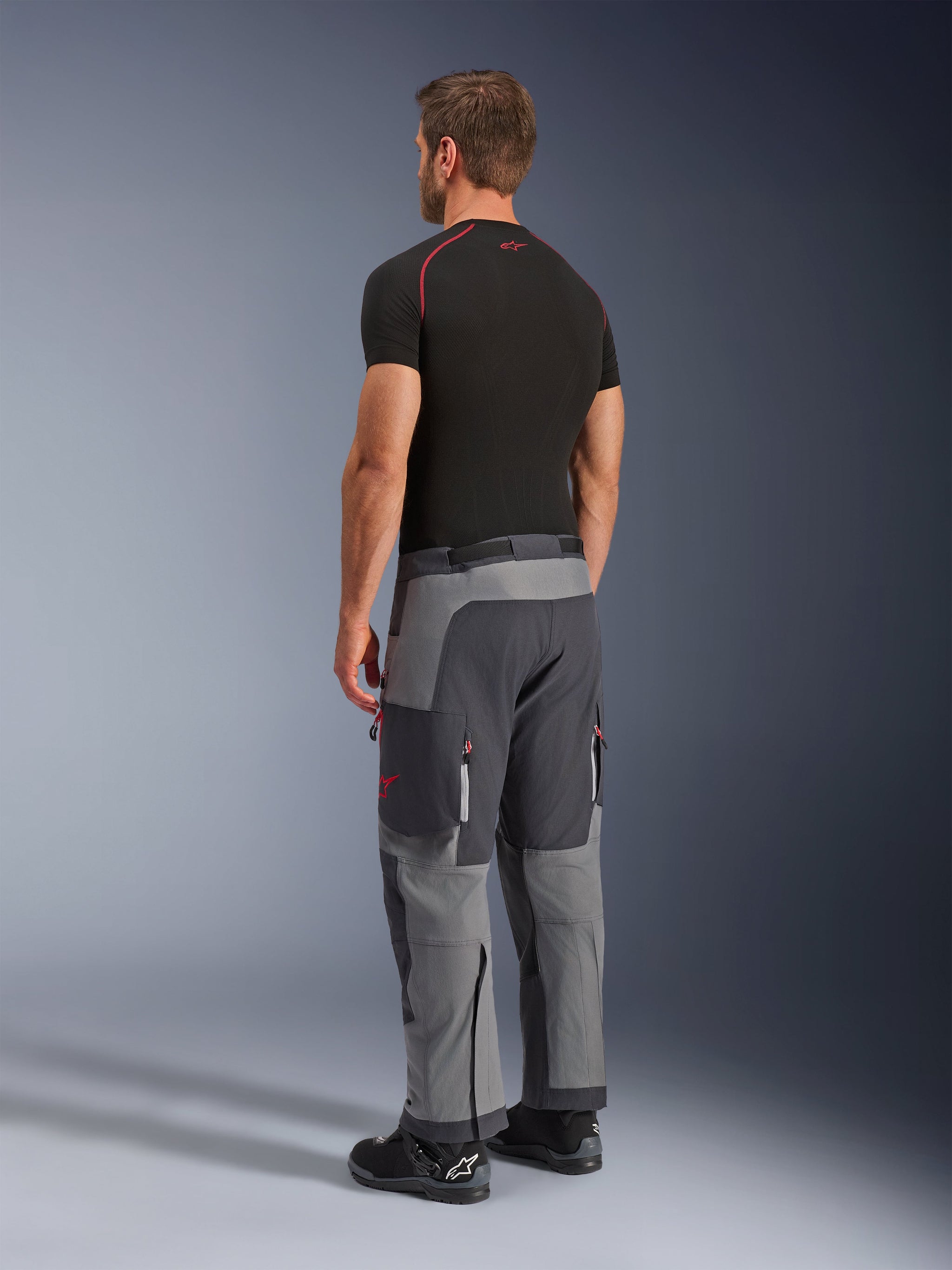 Alpinestars Maxdura Dual Pants, adventure motorbroek, donkergrijs en diepgrijs, gedragen door een model van achteren opzij gezien, met rode accenten op ritsen en logo, versterkte grijze panelen, cargozakken en over-de-laars pasvorm