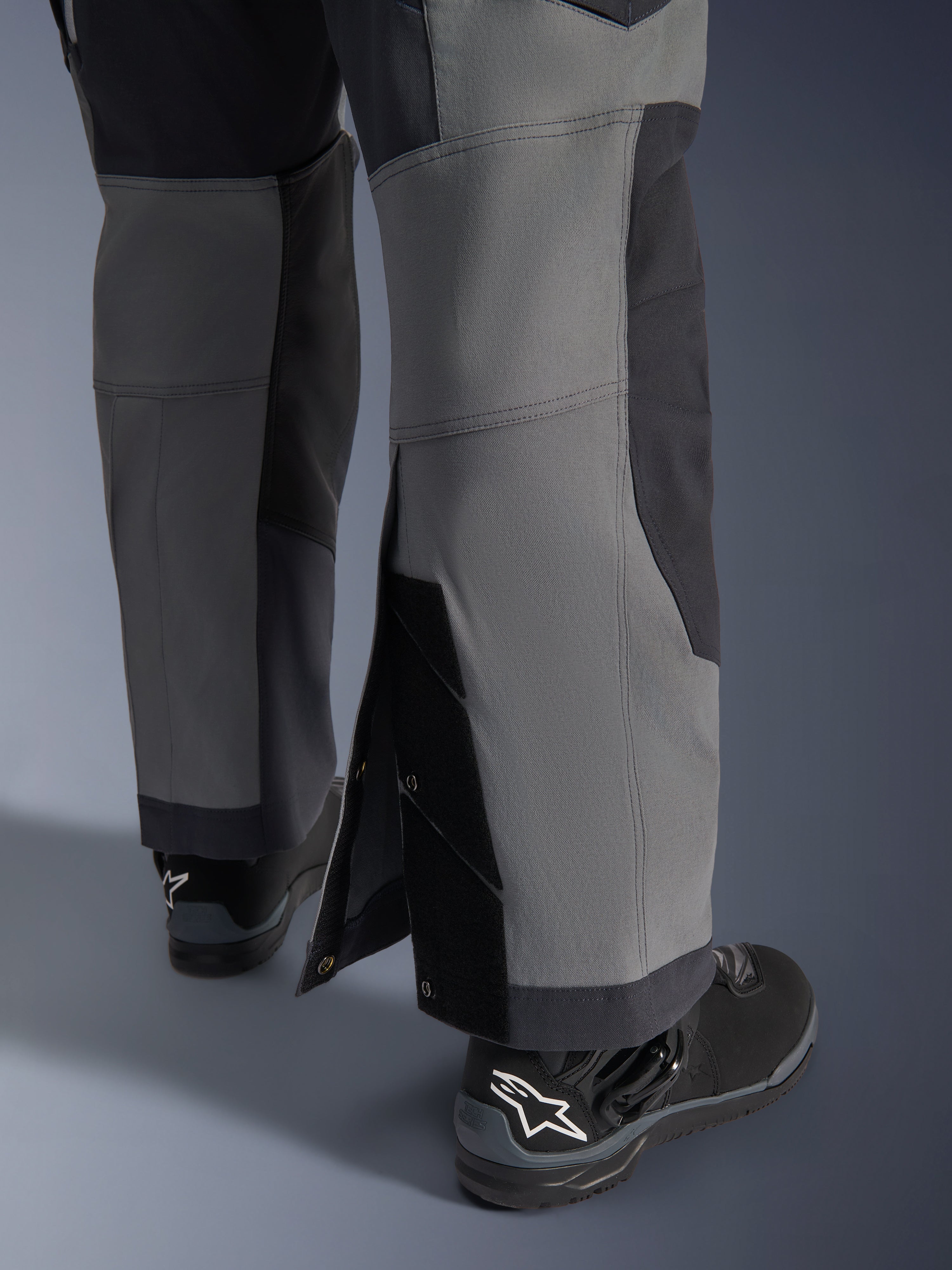 Alpinestars Maxdura Dual Pants, adventure motorbroek, donkergrijs en diepgrijs, close-up van de verstelbare over-de-laars broekzoom met drukknoopsluitingen en verstevigde panelen, gedragen met zwarte Alpinestars laarzen, robuust technisch ontwerp voor dual-sport ritten