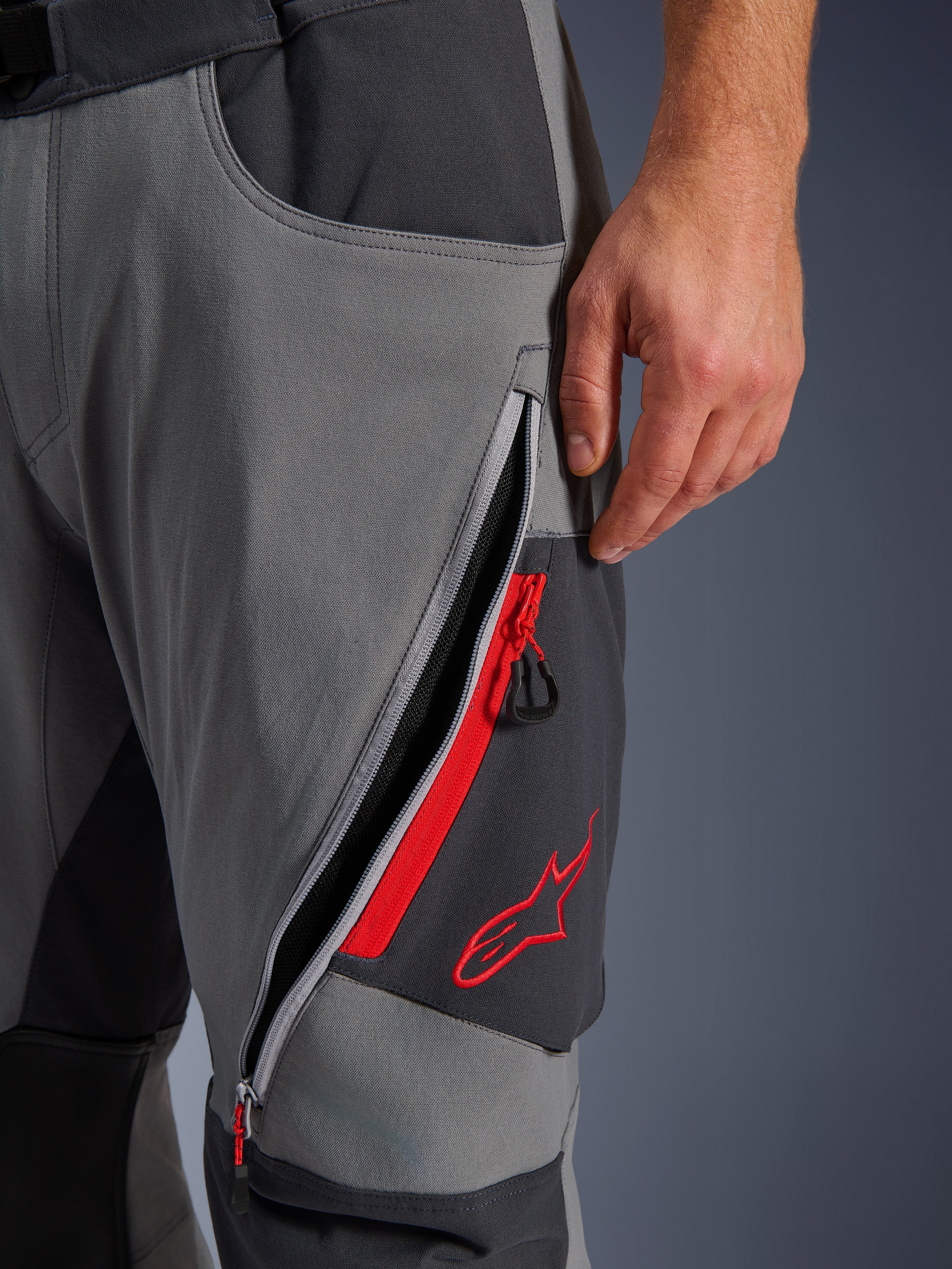 Alpinestars Maxdura Dual Pants, donkergrijze en diepgrijze adventure motorbroek, close-up van open ventilatieopening met rits op het bovenbeen met rode accenten en geborduurd rood sterlogo, technisch ontwerp van stretchstof