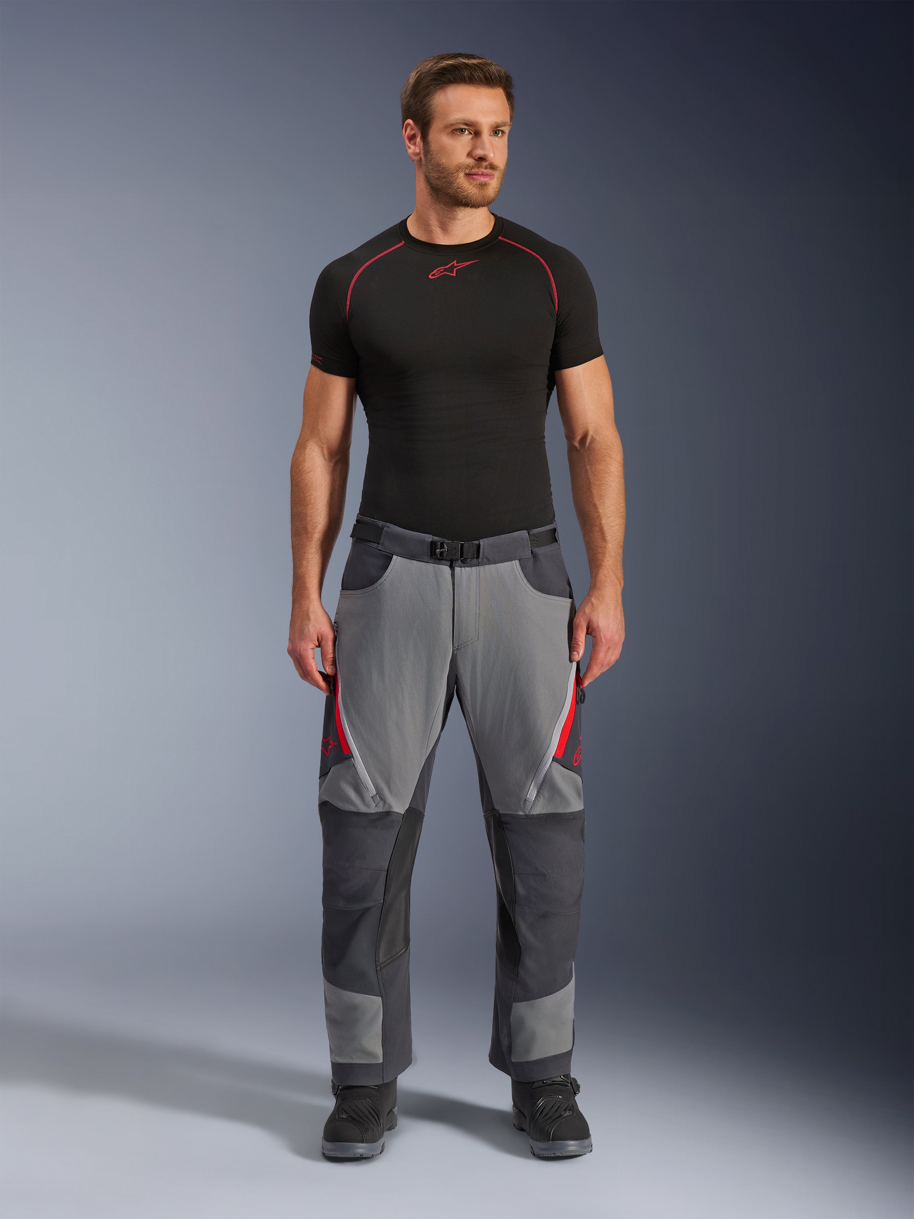 Alpinestars Maxdura Dual broek, motorbroek, donkergrijs en diepgrijs met rode accenten, vooraanzicht gedragen door een model met een zwarte Alpinestars base-layer en zwarte laarzen, voorzien van verstevigde panelen en ventilatieopeningen met rits op de bovenbenen, adventure-stijl motoruitrusting
