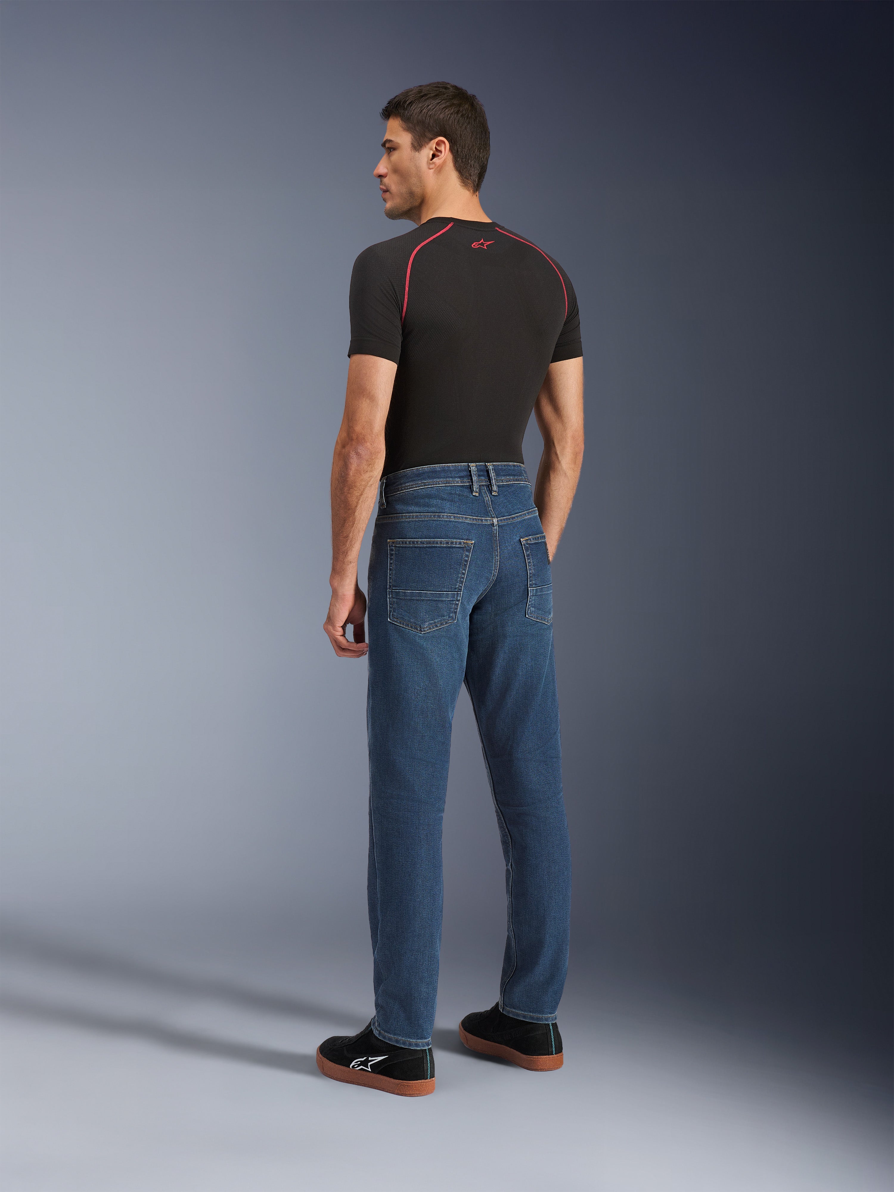 Alpinestars Flex-AST 5 Denim broek, Indigo Washed, stedelijke motorbroek met een toelopende pasvorm, van achteren getoond op een model met een zwart T-shirt en sneakers, voorzien van een strak vijfzakkenontwerp en monolaag stretchconstructie