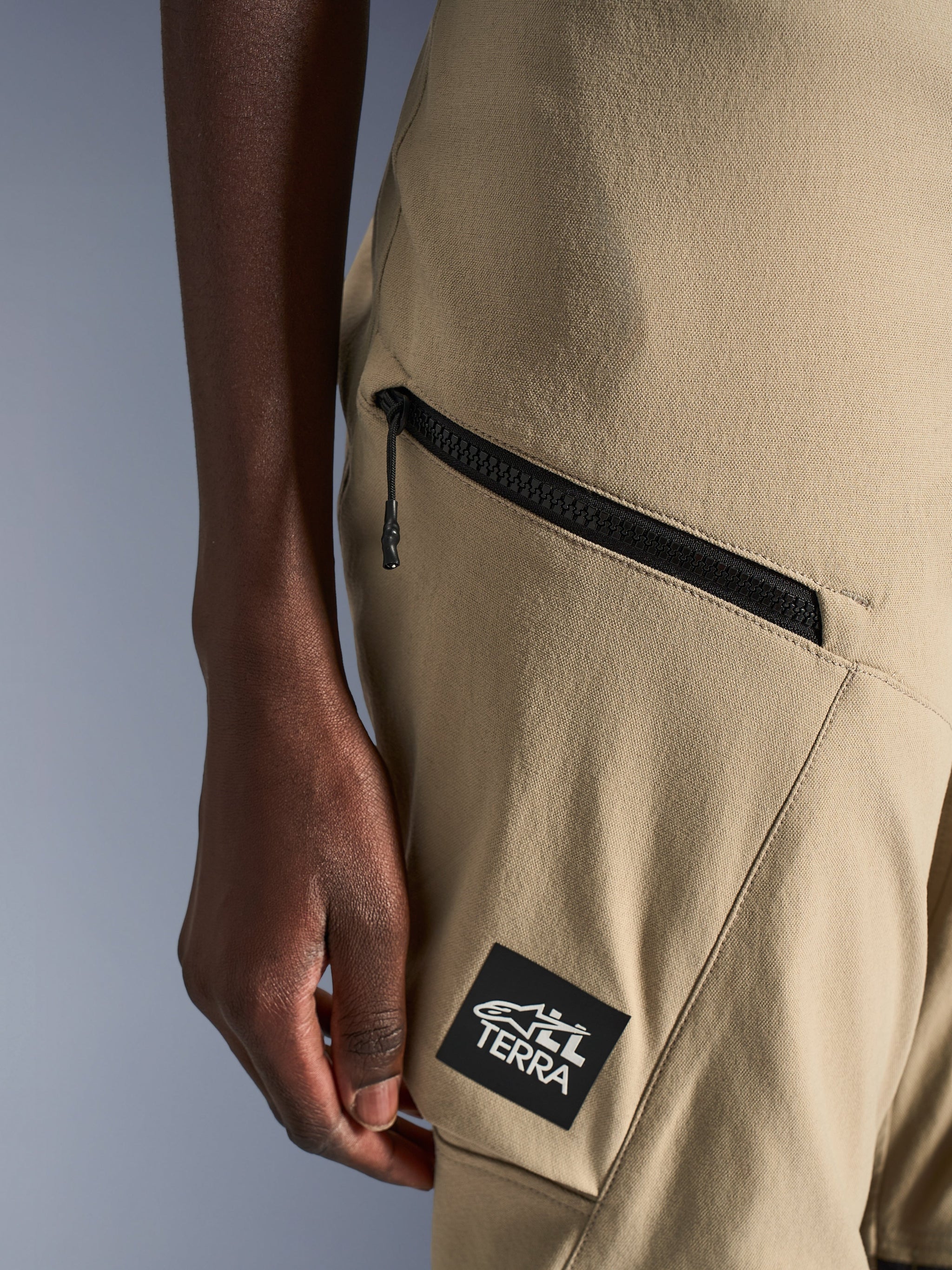 Alpinestars Stella Flex-AST Explorer broek, motorbroek voor dames, zandkleur, close-up van een cargozak met ritssluiting en zwarte ALL TERRA-logopatch, duurzame ripstop-textielconstructie voor adventure touring