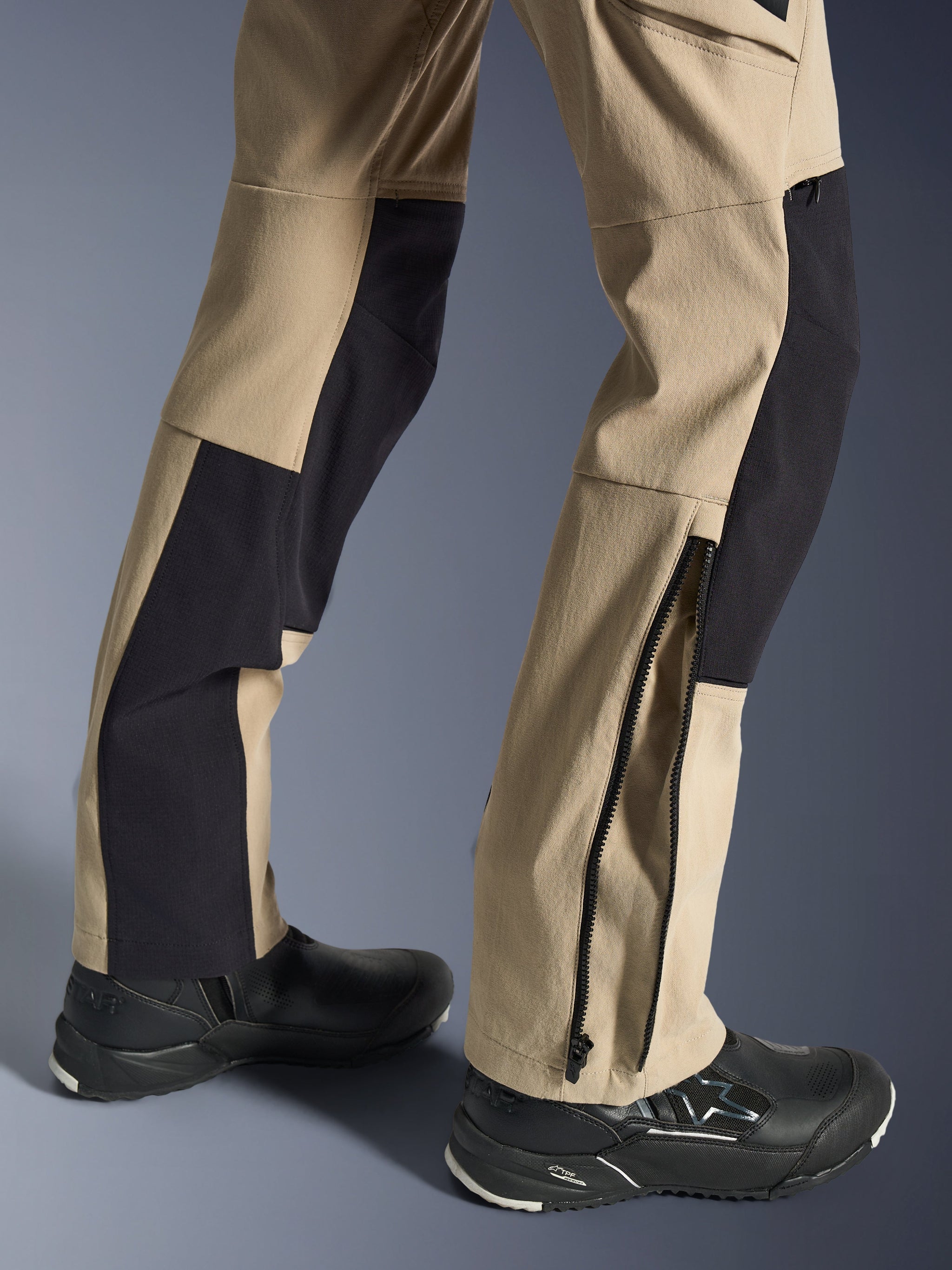 Alpinestars Stella Flex-AST Explorer broek, dames motorbroek, zandkleur en zwart, close-up van de onderbenen met een ritssluiting bij de zoom voor pasvorm over laarzen, gedragen met zwarte Alpinestars motorschoenen, voorzien van duurzame ripstop en ademende stretchstof panelen.