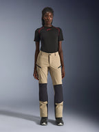 Alpinestars Stella Flex-AST Explorer damesmotorbroek, beige met zwarte panelen, gedragen door een model met een zwart shirt met korte mouwen en zwarte laarzen, voorzien van verstevigde kniepanelen, cargozakken met ritssluiting en ademende ripstop-stof voor adventure-ritten in warm weer.