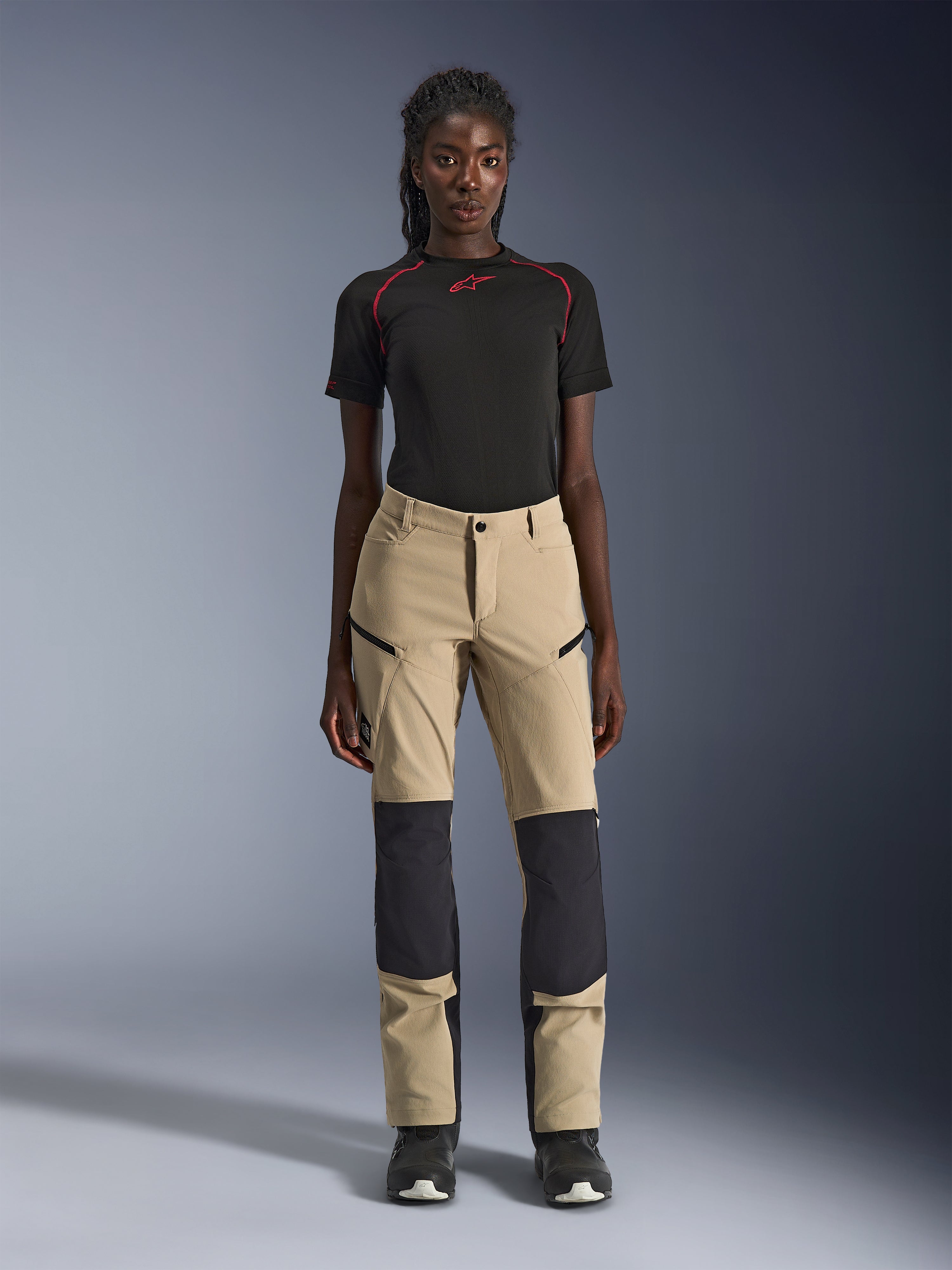 Alpinestars Stella Flex-AST Explorer damesmotorbroek, beige met zwarte panelen, gedragen door een model met een zwart shirt met korte mouwen en zwarte laarzen, voorzien van verstevigde kniepanelen, cargozakken met ritssluiting en ademende ripstop-stof voor adventure-ritten in warm weer.